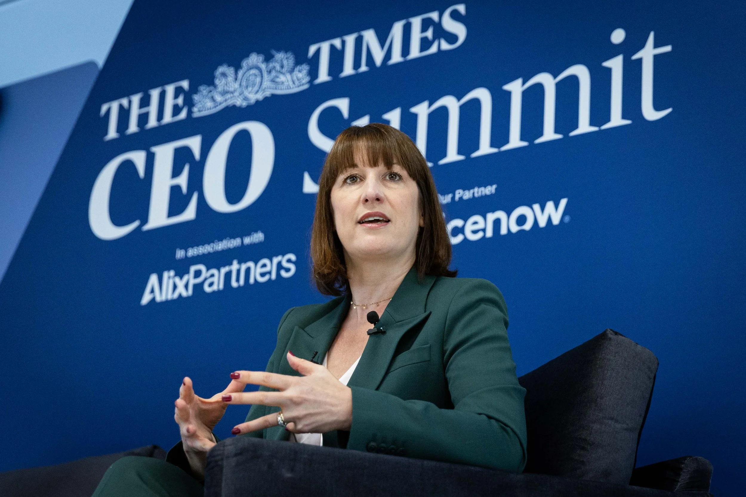 British Chancellor Rachel Reeves speaking at an event, seated in front of a blue backdrop with white text that reads 'The Times CEO Summit' and 'In association with AlixPartners.' She is wearing a dark green blazer and gesturing with her hands.