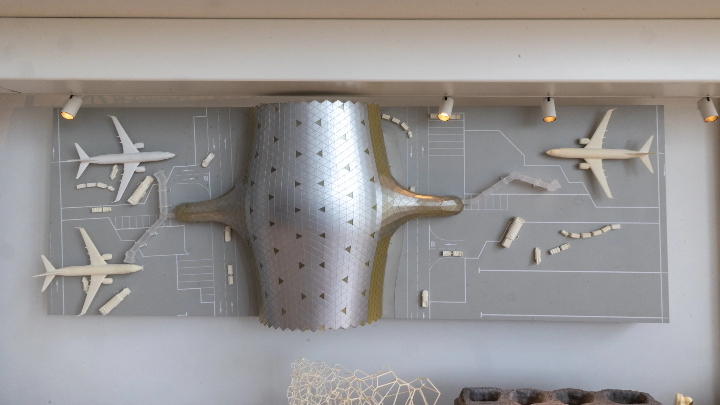 A miniature architectural model of a modern building with a unique, curved, pyramidal shape, surrounded by toy airplanes and vehicles on a gray base with white lines and parking spaces.