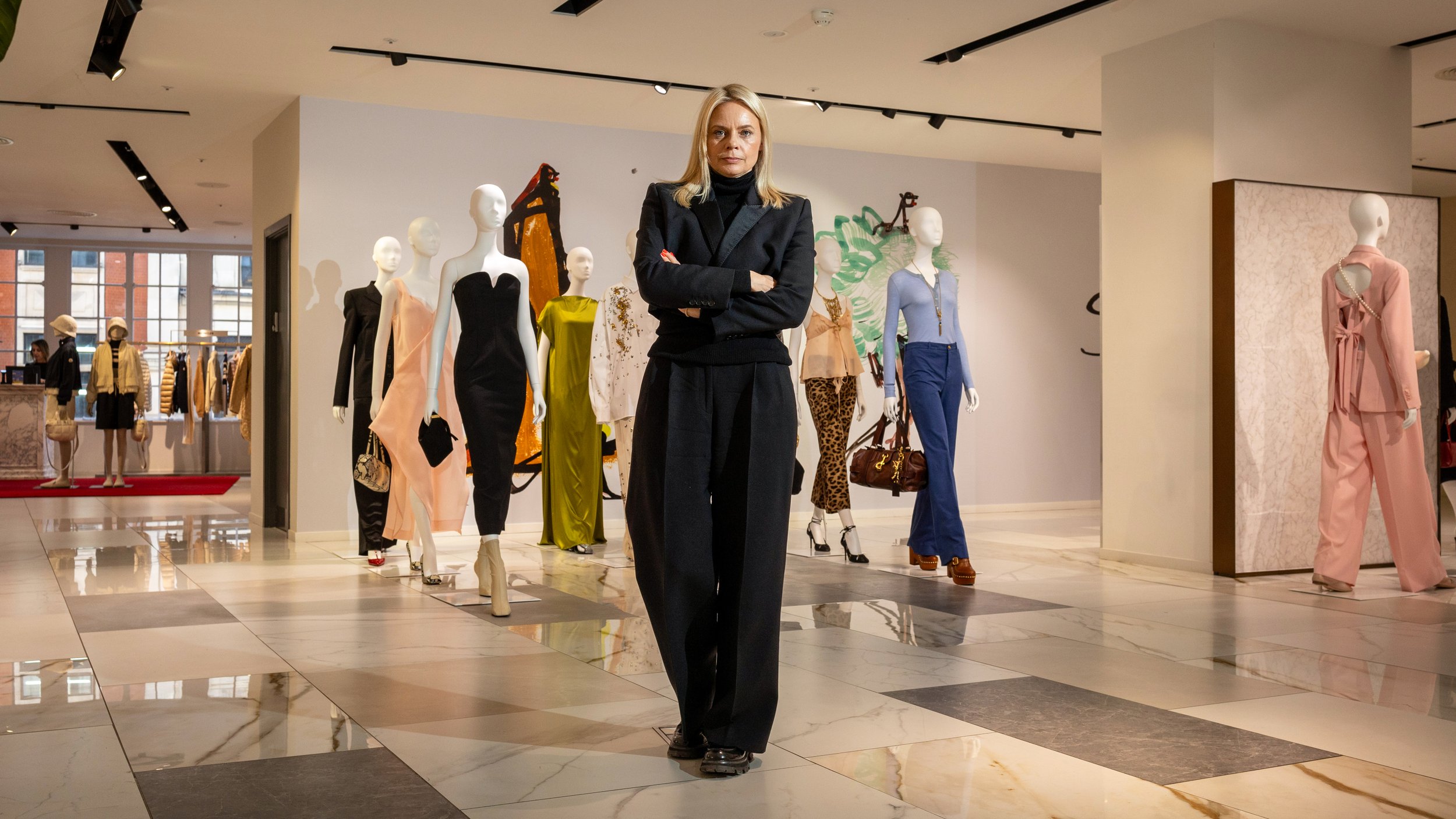 CEO Julia Goddard wears black pants and a black blazer walks through a clothing store with mannequins dressed in colorful outfits around her.