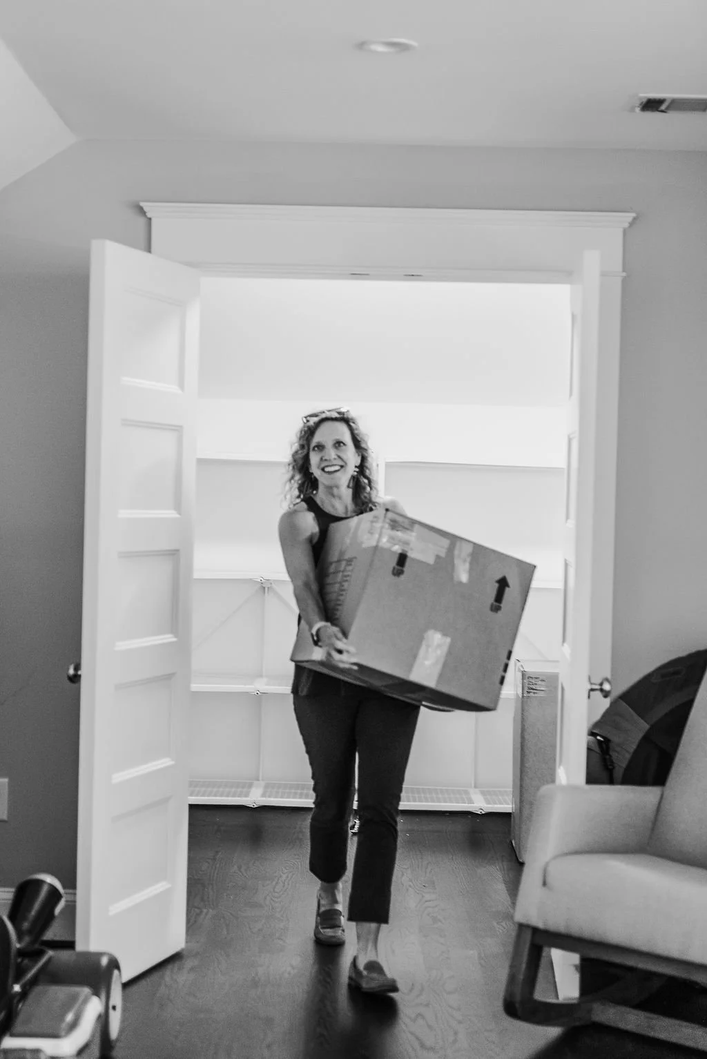 A woman smiling and carrying a large box enters a room through open double doors in a home, with an armchair on the right and a backpack in the background.