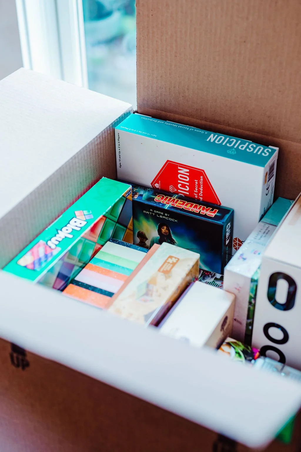A cardboard box filled with various board games and puzzle boxes, including Rush Hour and Pandemic, inside a well-lit room near a window.