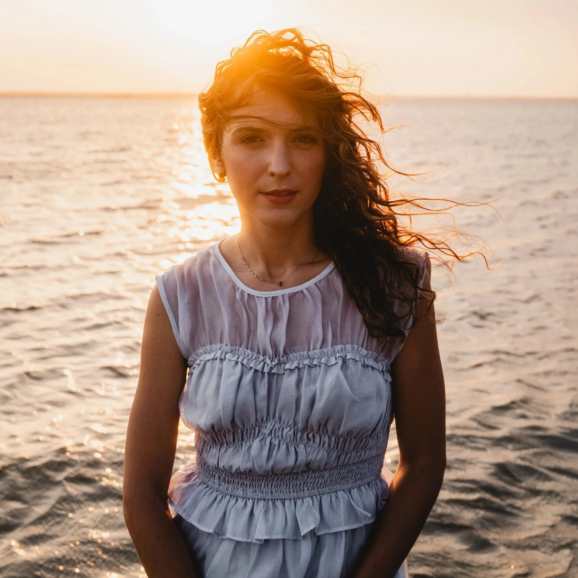 A woman with curly hair and earrings stands on a beach at sunset, facing the camera with the ocean and a bright sunset in the background.