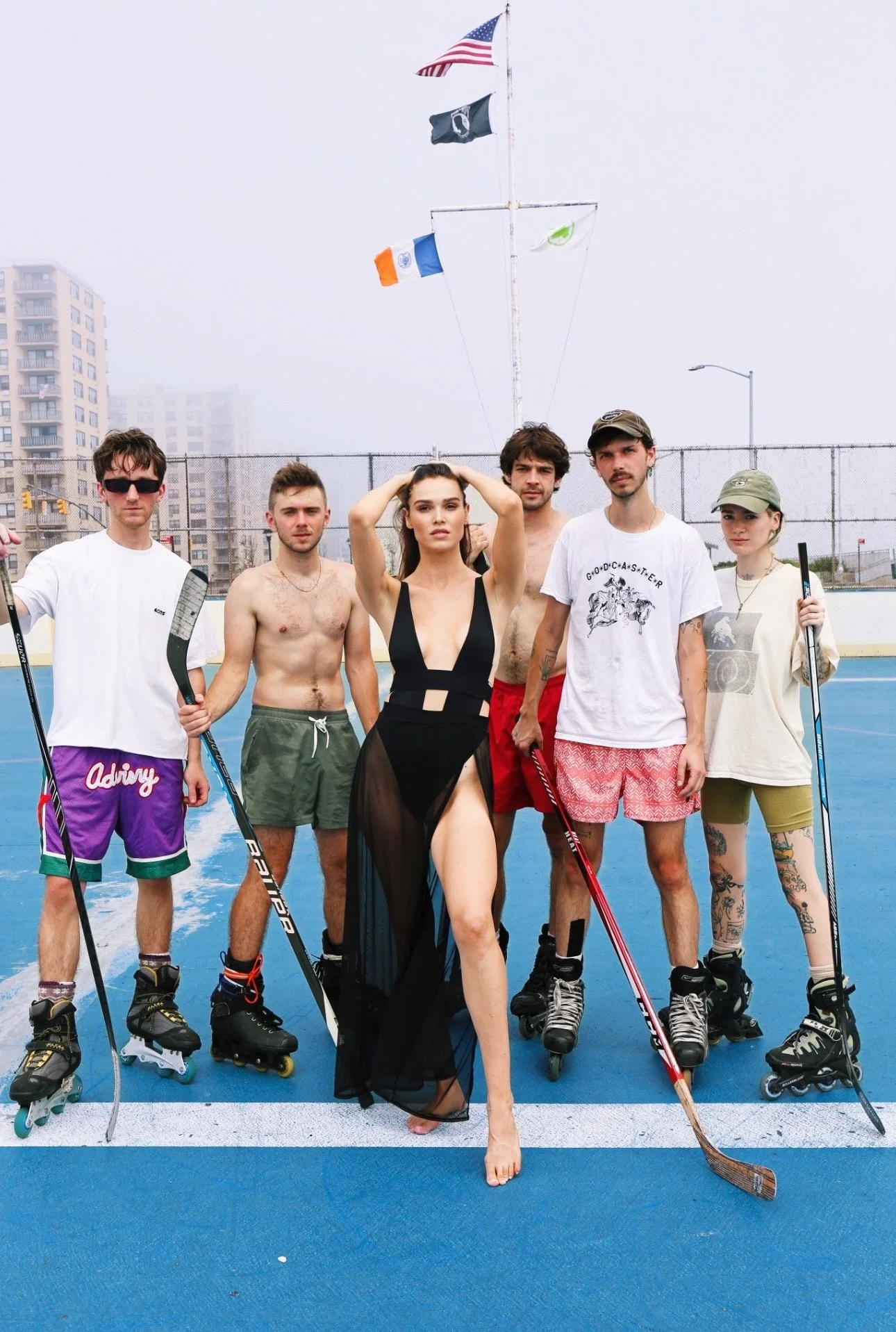 A group of seven young adults on roller skates posing on an outdoor sports court with American and other flags in the background. One woman is in the center wearing a black sheer dress, and the others are in casual summer or sportswear.
