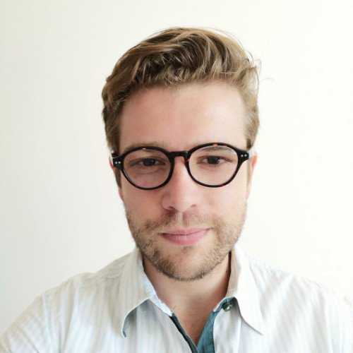 Primer plano de un hombre con gafas de montura negra, cabello castaño claro peinado hacia atrás, barba de unos días, con camisa blanca y fondo neutro.