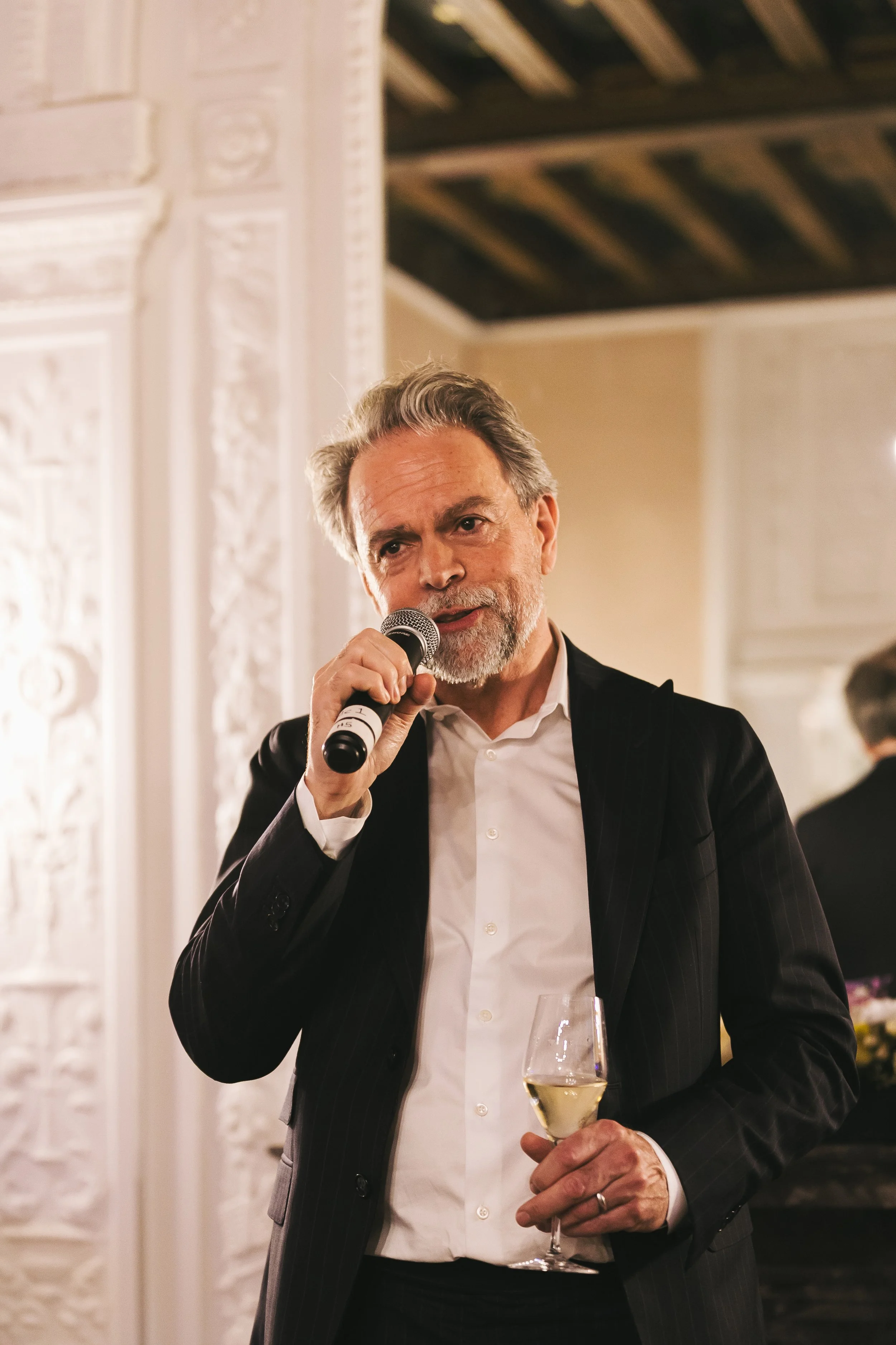 NAHC A man with gray hair and a beard wearing a dark pinstripe suit and white shirt, holding a microphone and a glass of white wine, appears to be speaking at an event in an elegant room with ornate wall decorations.