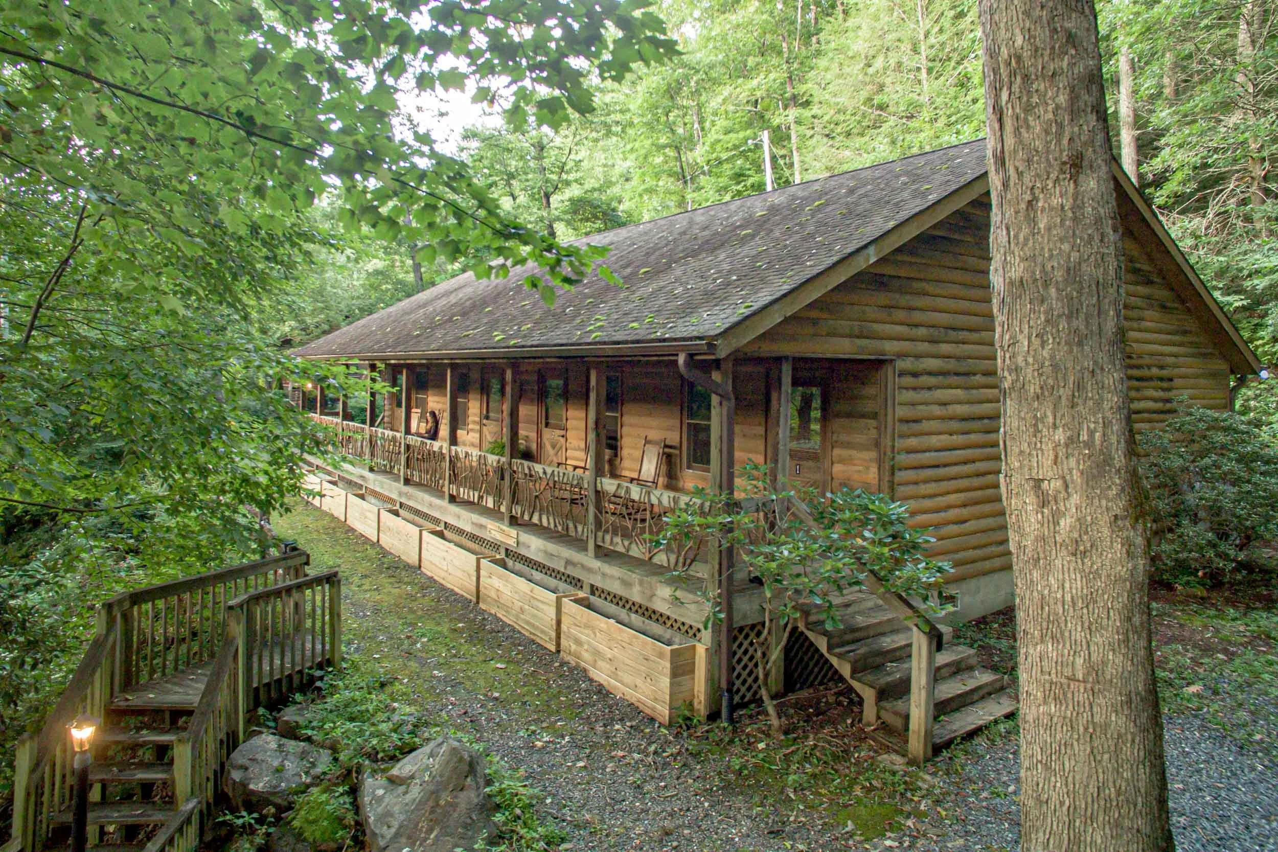 Wooden cabin in a forested area with a porch and steps leading up to it. The cabin is made of logs and has a pitched roof. Surrounding trees and greenery.