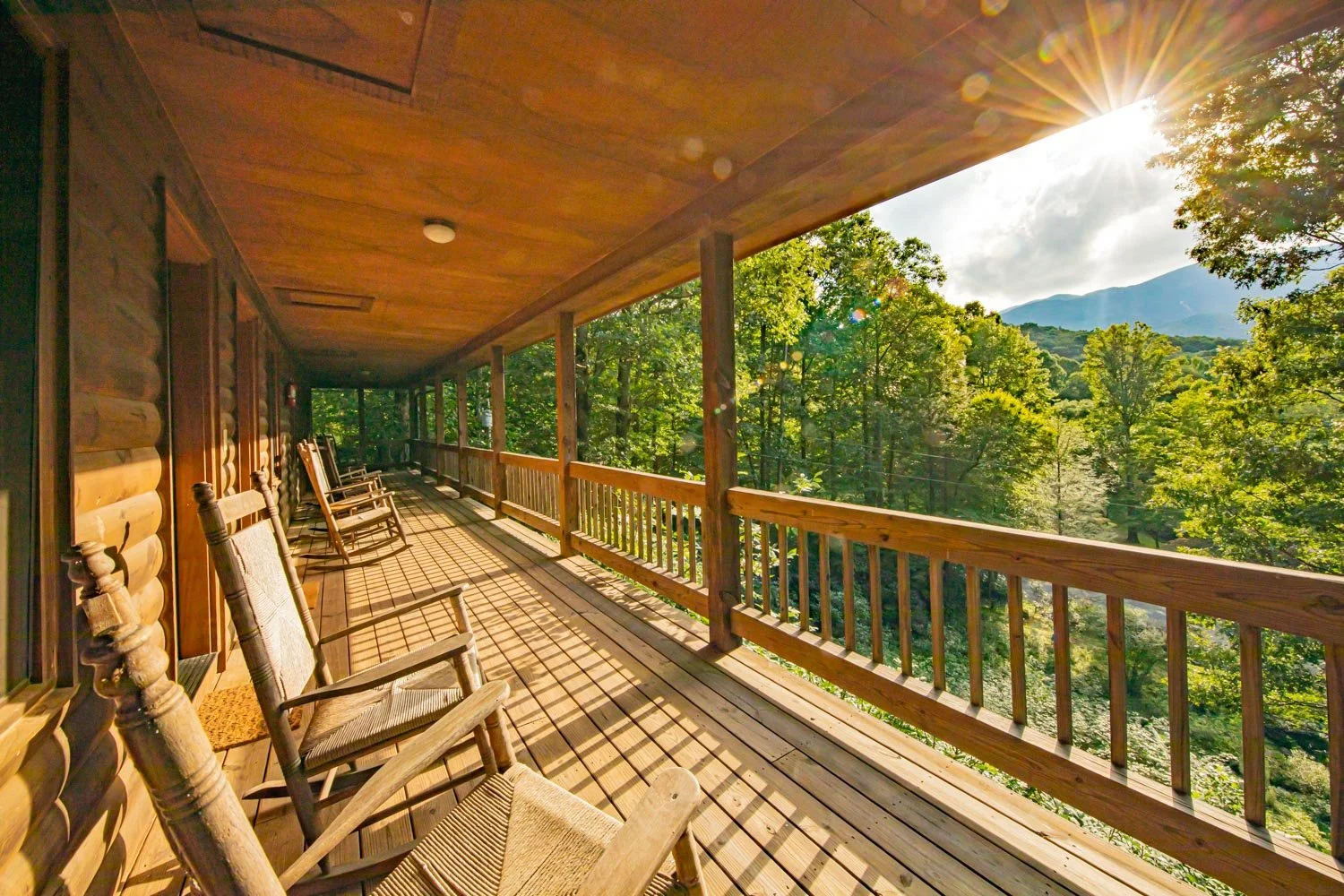 Wooden porch with rocking chairs overlooking a forested landscape and distant mountains, sun shining through trees.