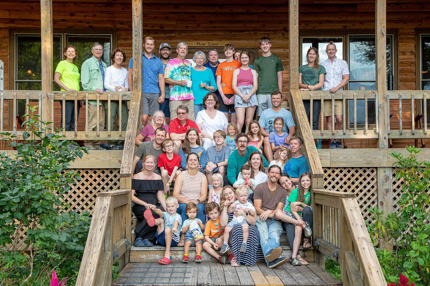 A large group of people, including children and adults, posing for a photo on the steps and porch of a wooden lodge or cabin.