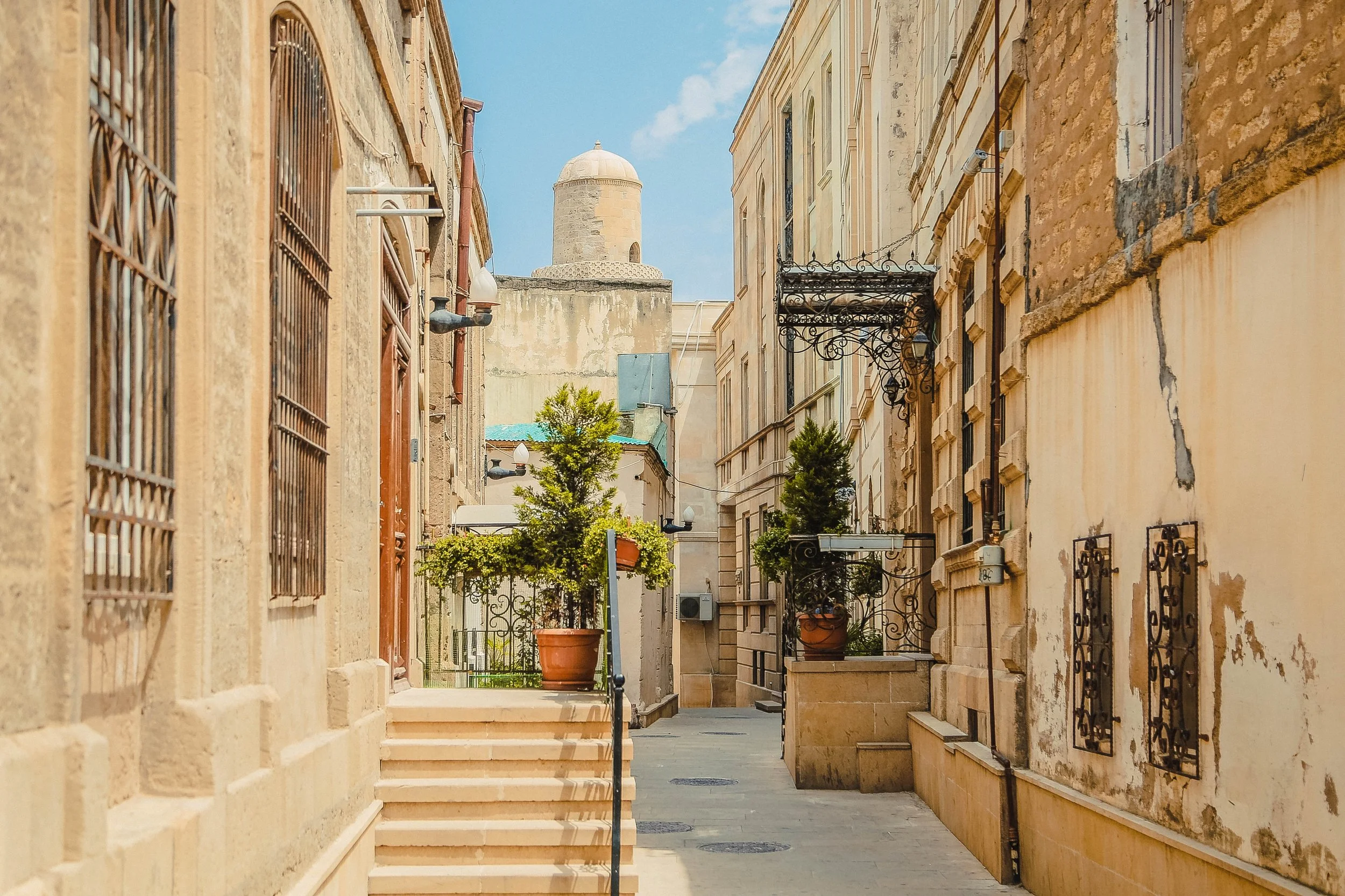 A narrow street in an old city with closely built stone buildings, potted plants, and a rooftop tower in the background under a partly cloudy sky.
