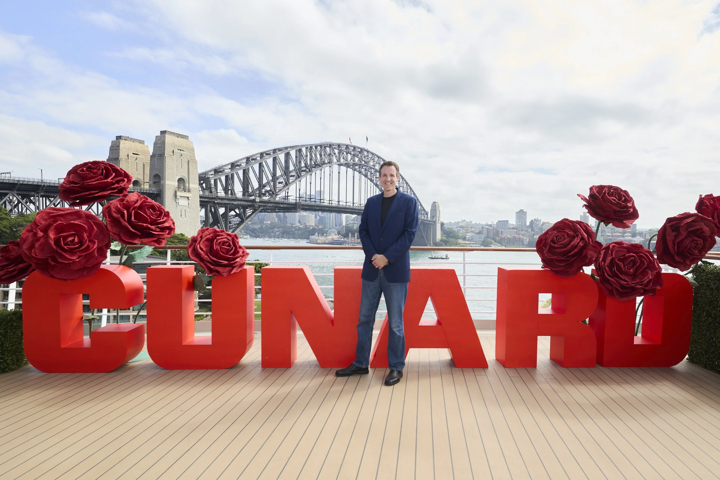 A man in a blue blazer and jeans standing smiling in front of large red letters spelling 'LOVE' with red roses attached to the letters, on a deck near water with the Sydney Harbour Bridge and city skyline in the background, under a partly cloudy sky.