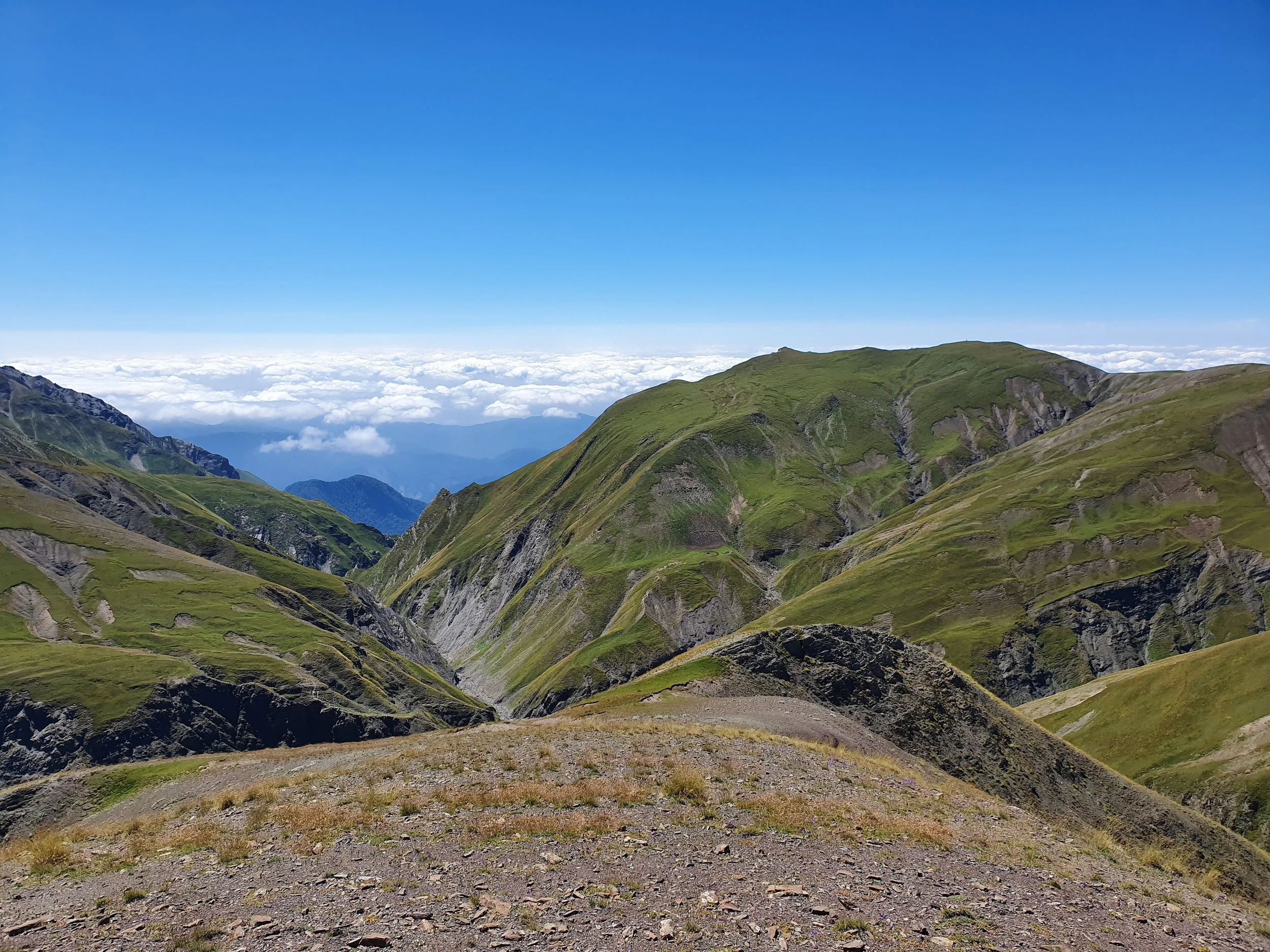 Scenic view of green mountains with patches of rocky terrain beneath a clear blue sky and clouds in the background.