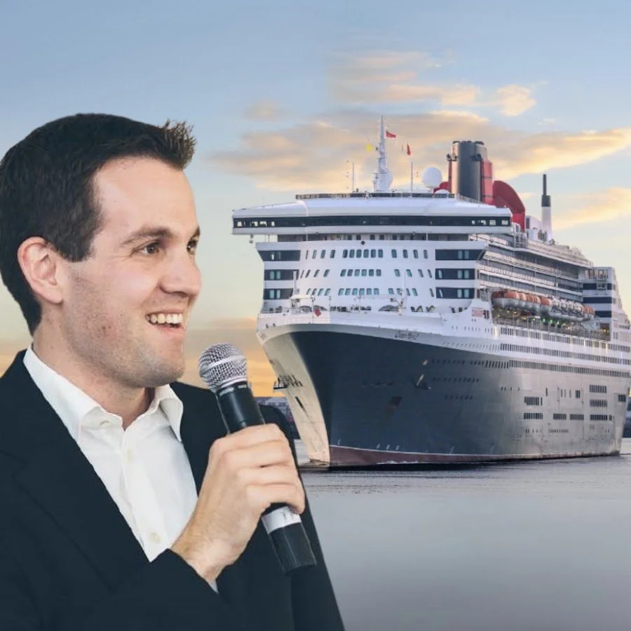 Man in a suit holding a microphone speaking with a cruise ship in the background during sunset.