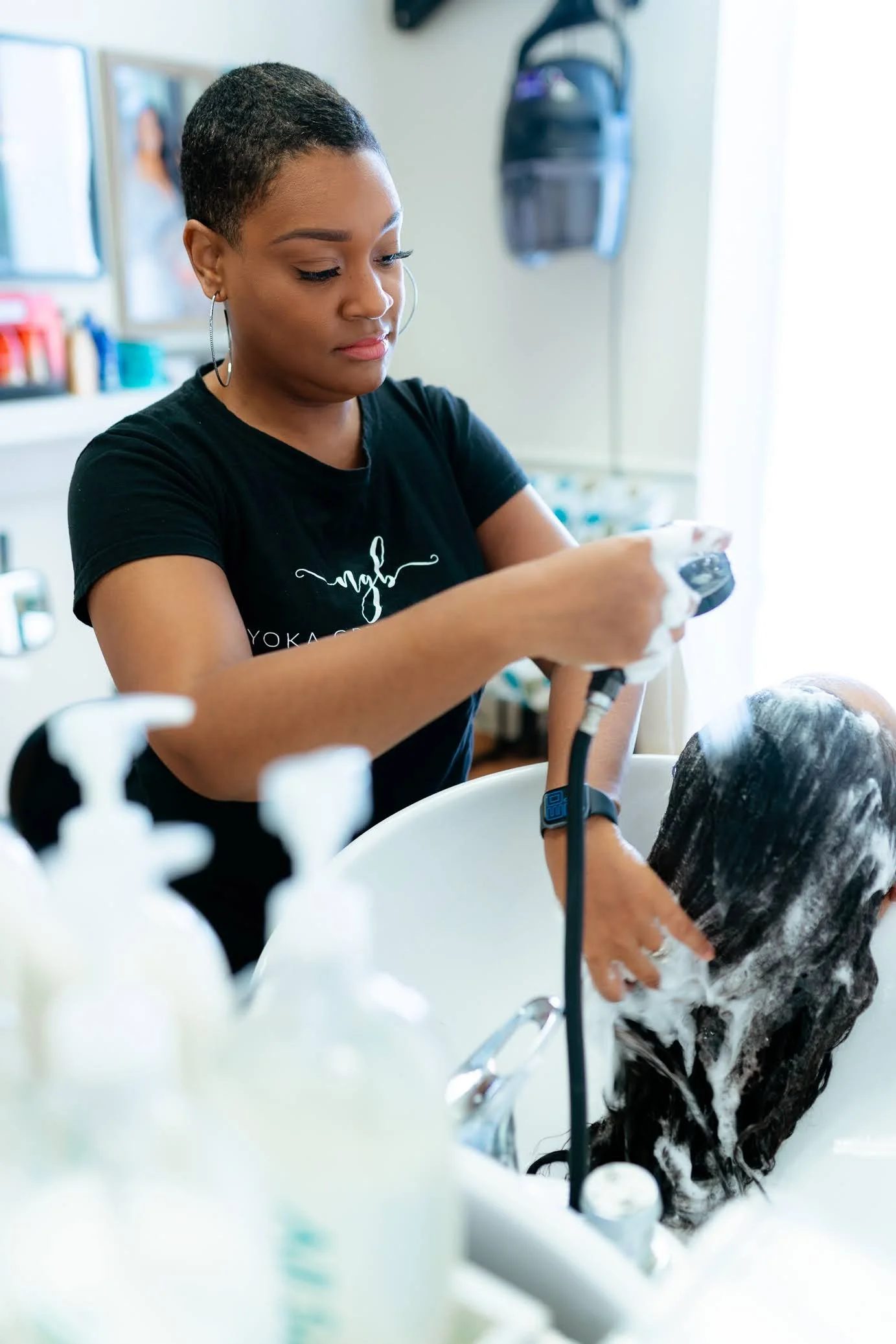 An African American hairstylist in a black short sleeve shirt is rinsing shampoo from her clients hair.
