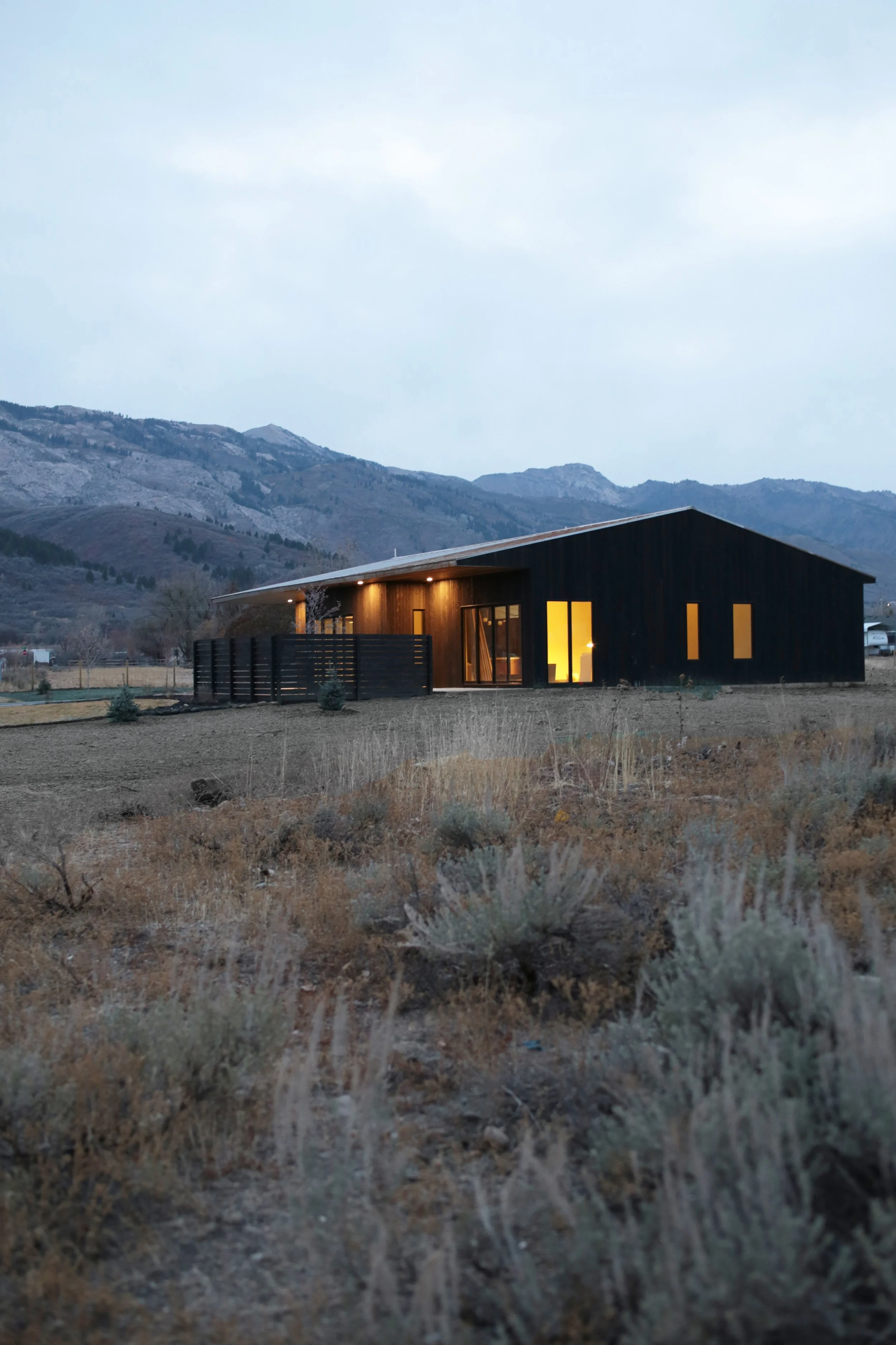 Modern black house with illuminated windows in a rural landscape with dry grass and mountains in the background during dusk.