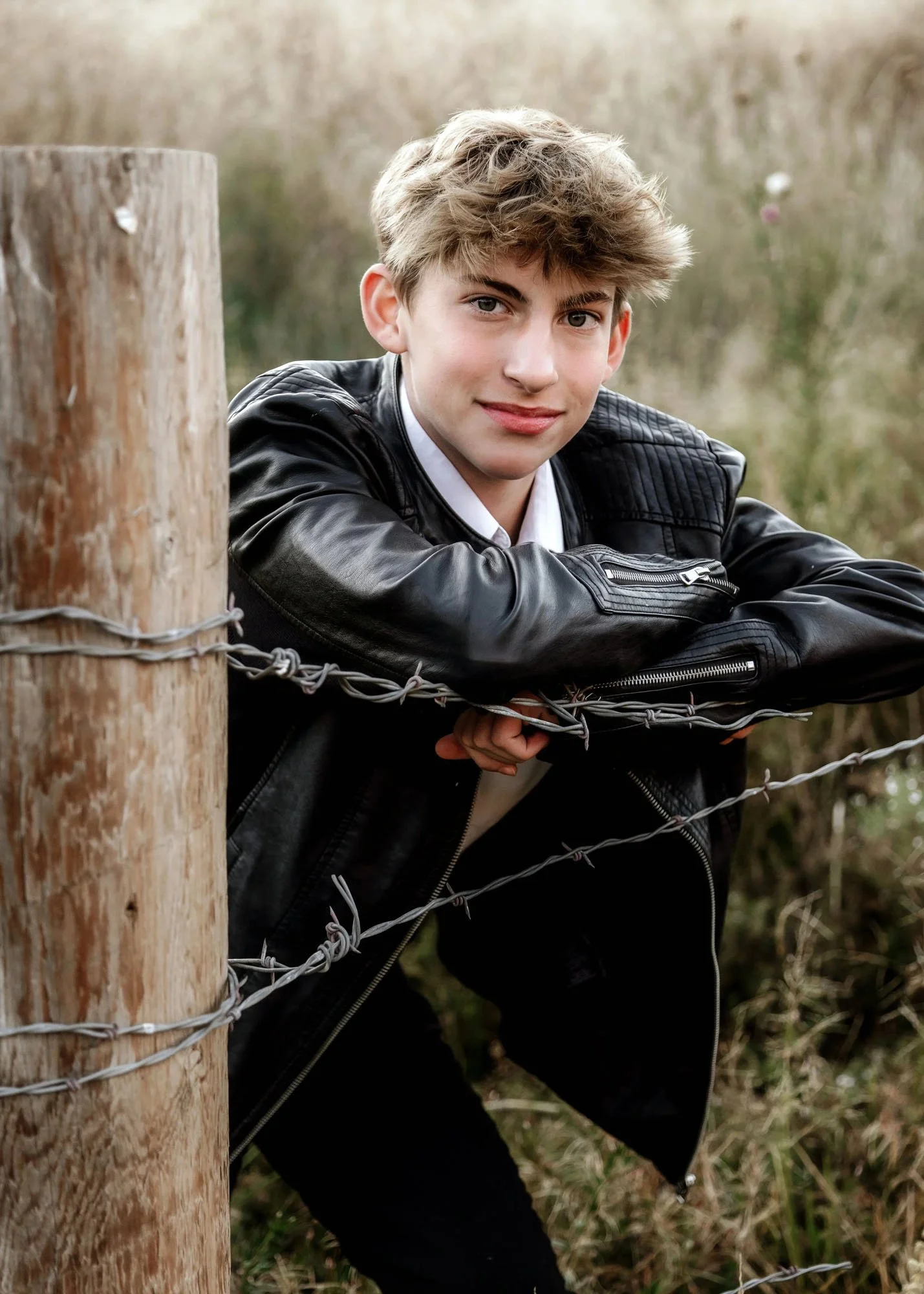 Teen boy in black leather jacket leaning on barbed wire fence looking at camera