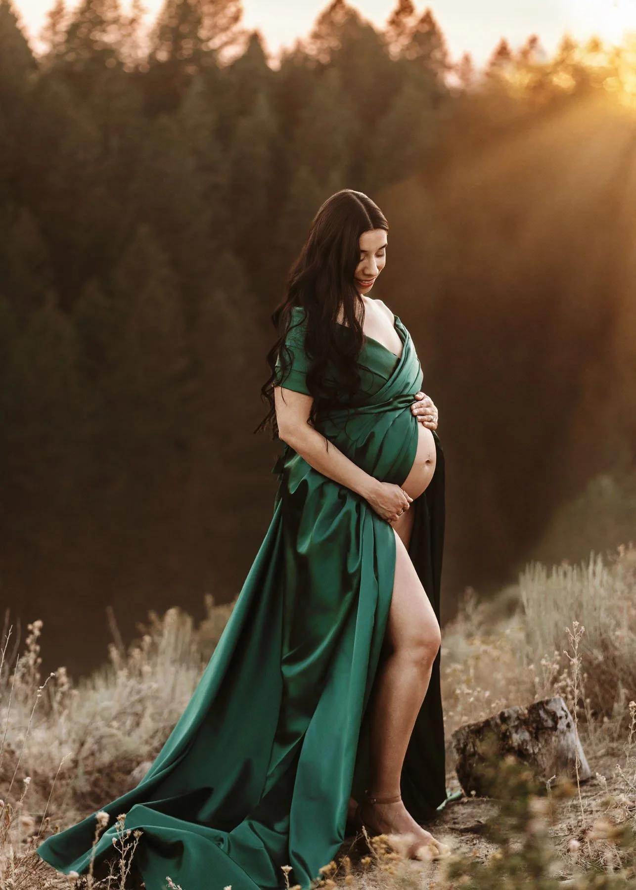 Mama in green draped dress showing baby bump in front of pine trees in mountains