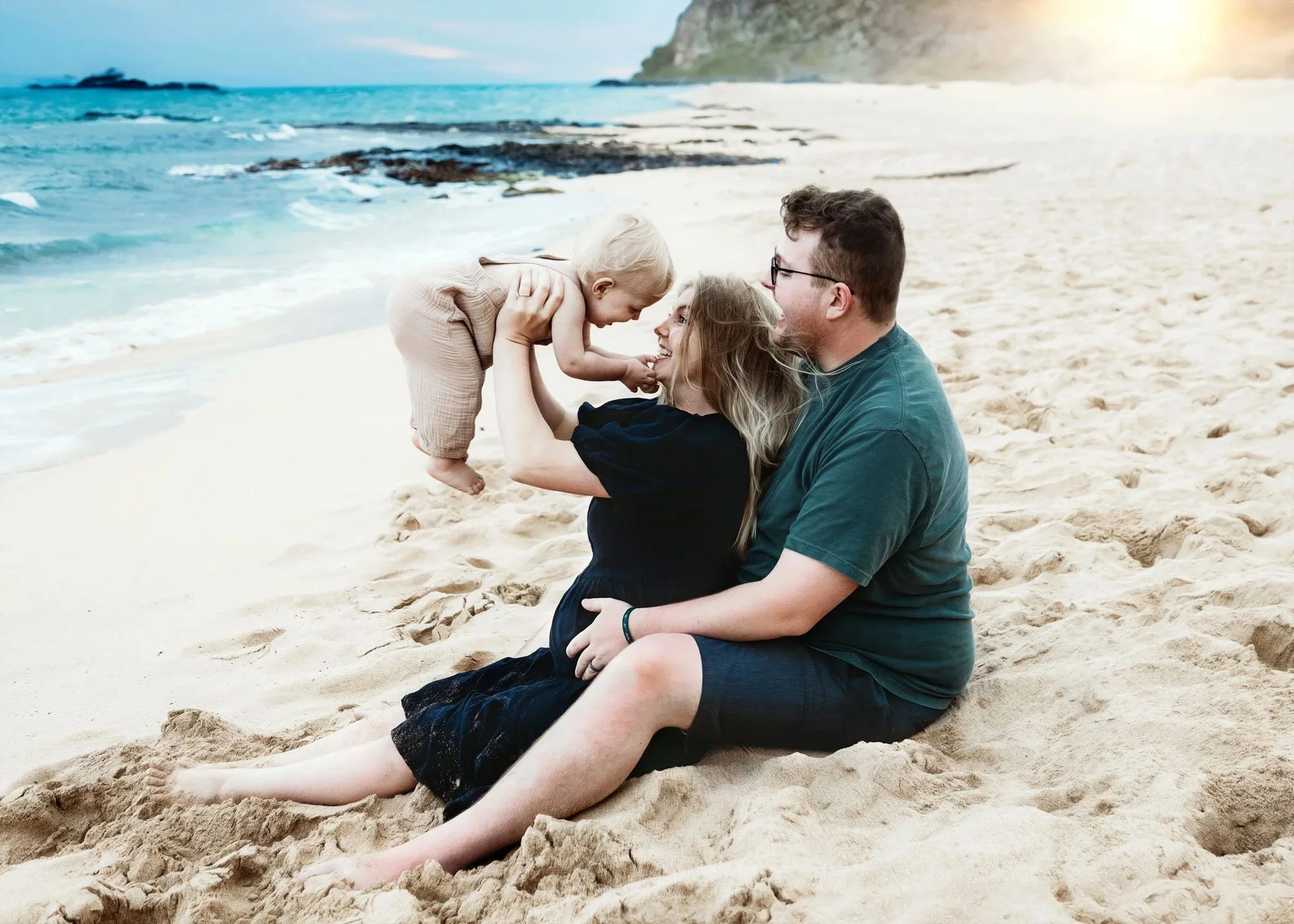 Couple sitting on the beach tossing toddler in the air, mama in black dress with baby bump visible