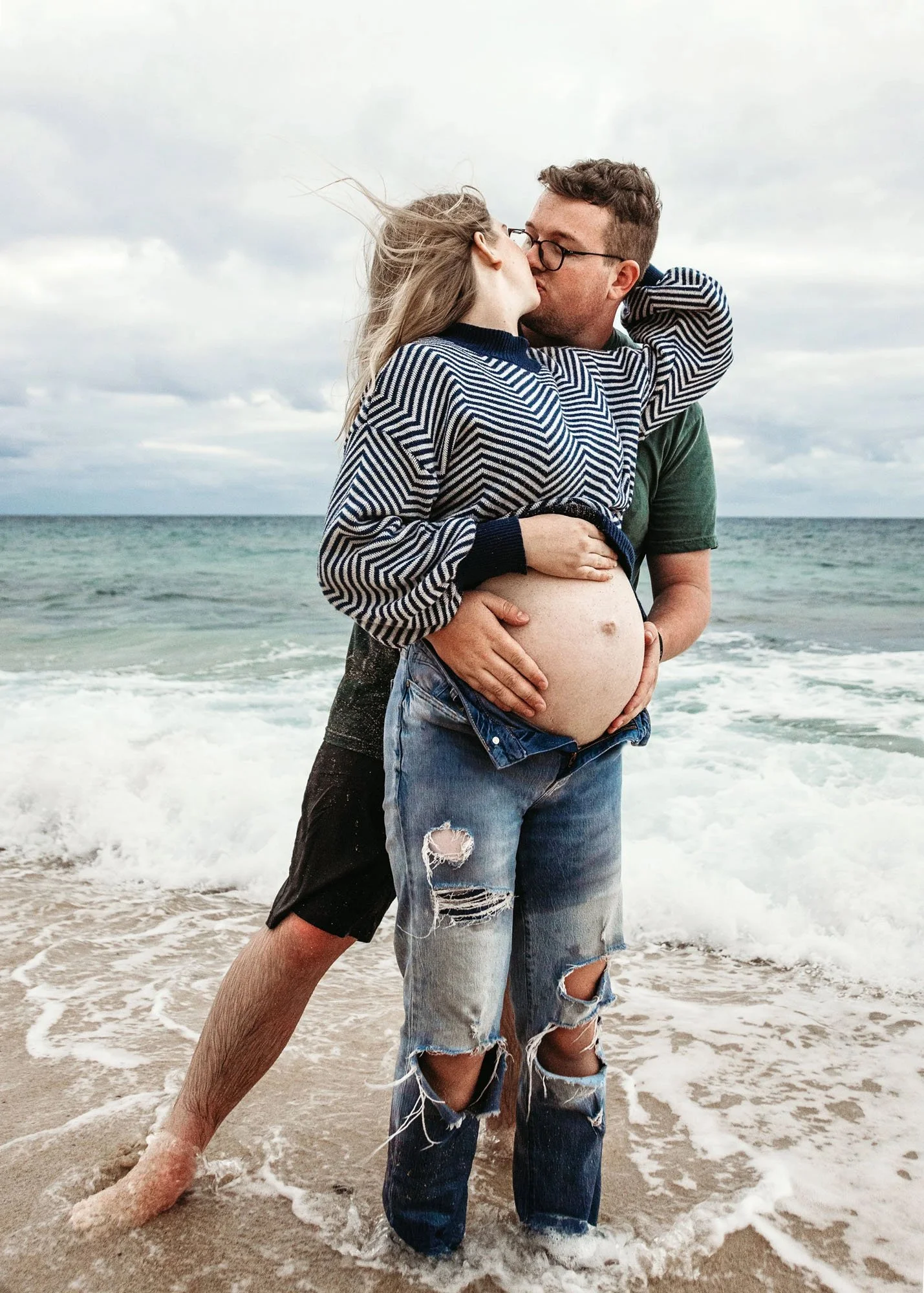 Pregnant mama in black dress holding toddler with one arm and supporting baby bump in front of the ocean