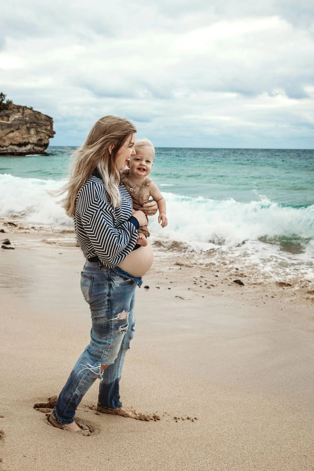 Pregnant mama holding toddler in one arm and baby bump in the other with ocean behind her