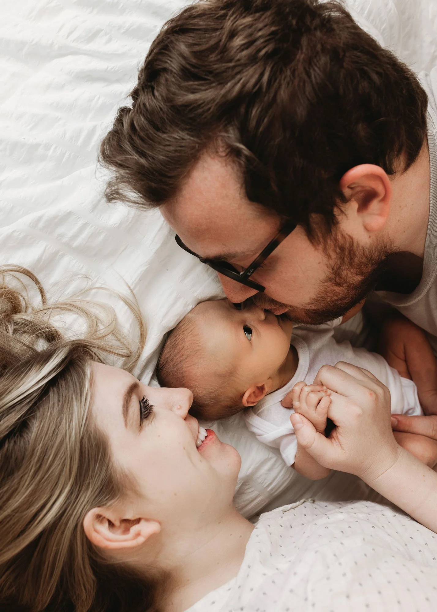 Family laying on bed with newborn in the middle and father kissing baby
