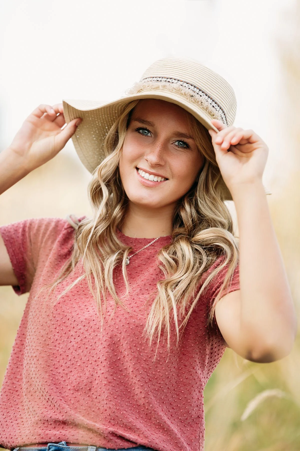 Senior girl smiling, holding her hat, wearing pink shirt in outdoor portrait