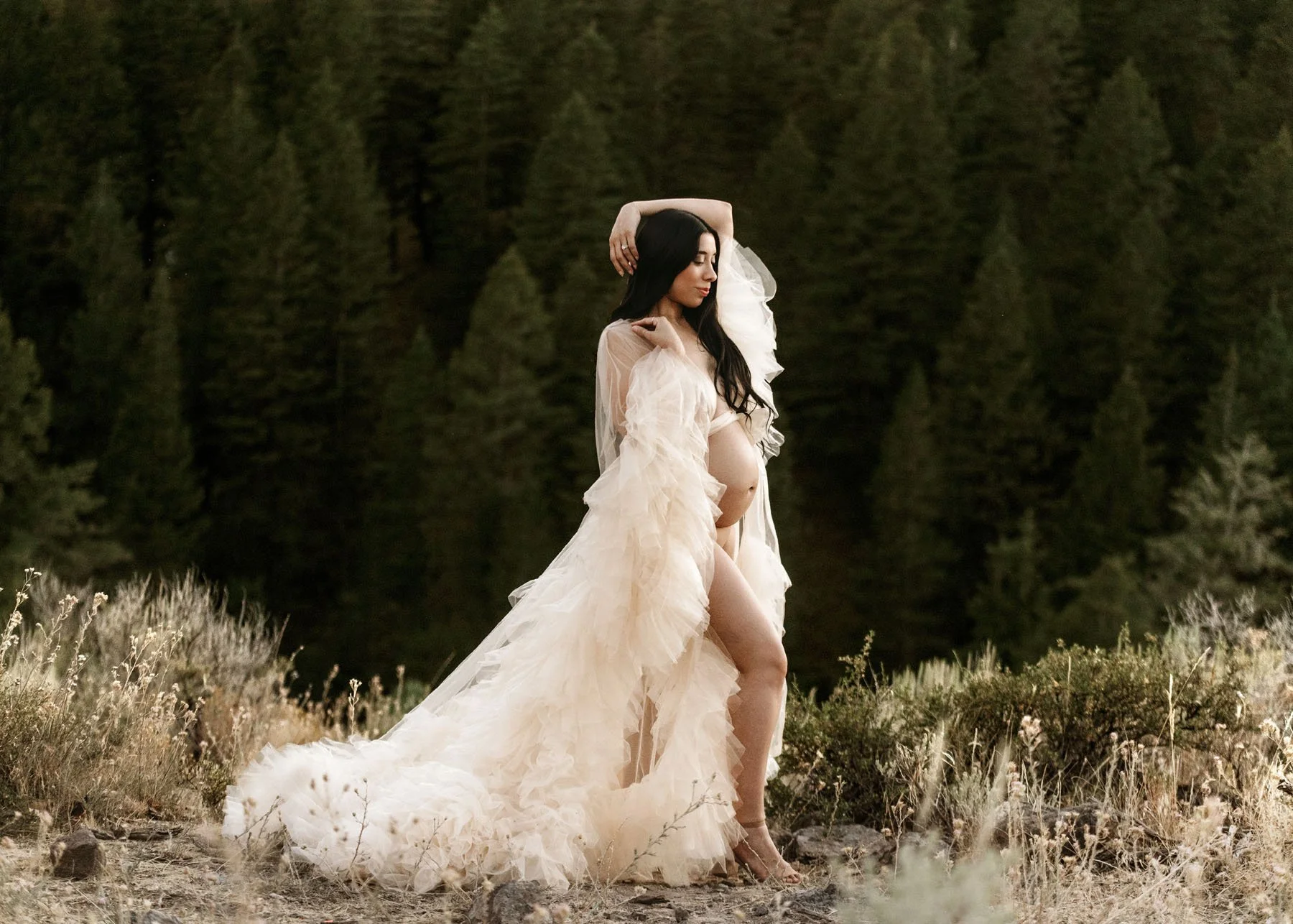 Mama in neutral fluffy dress showing baby bump in front of pine trees in mountains
