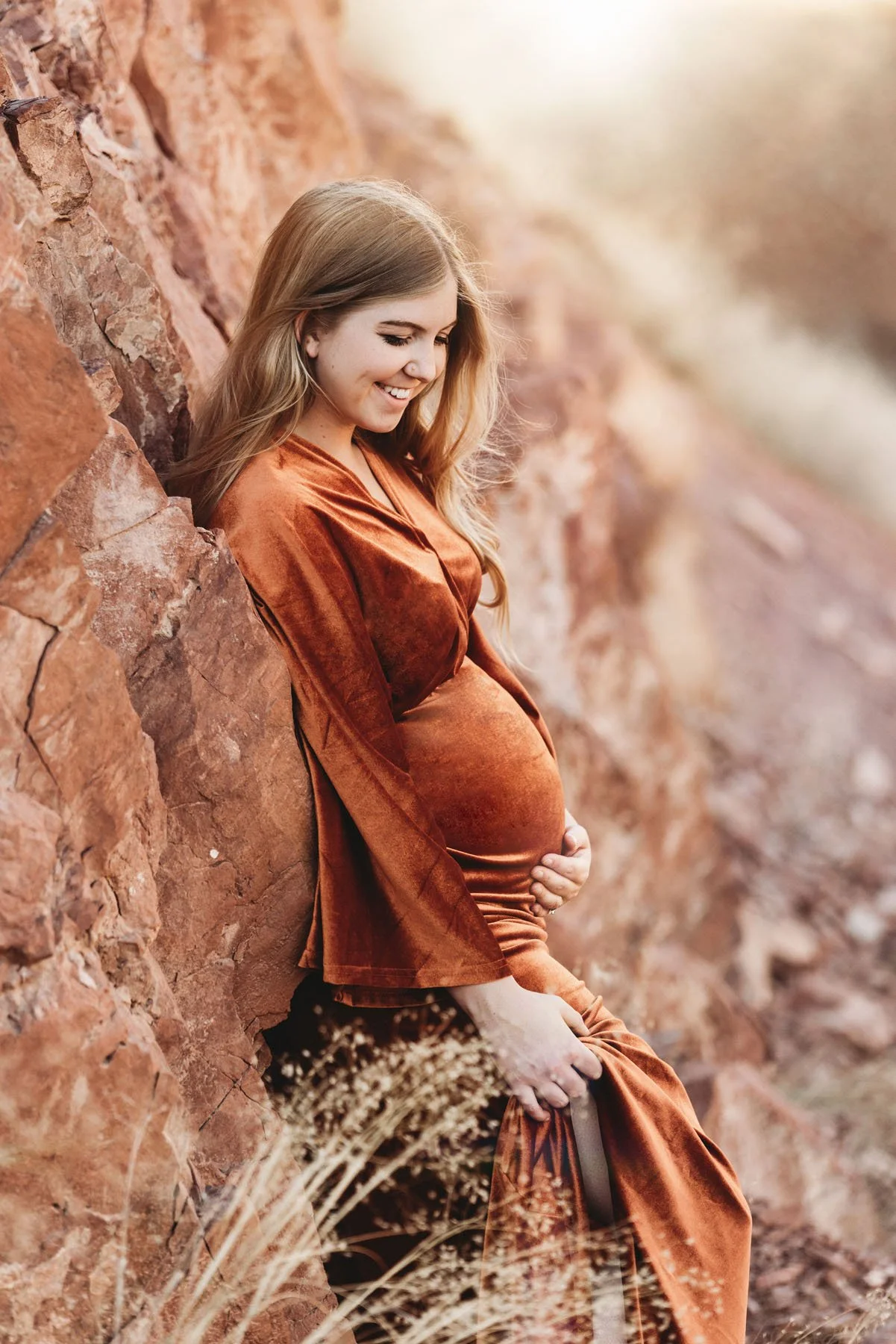 Mama in orange dress leaning against red rocks, baby bump visible