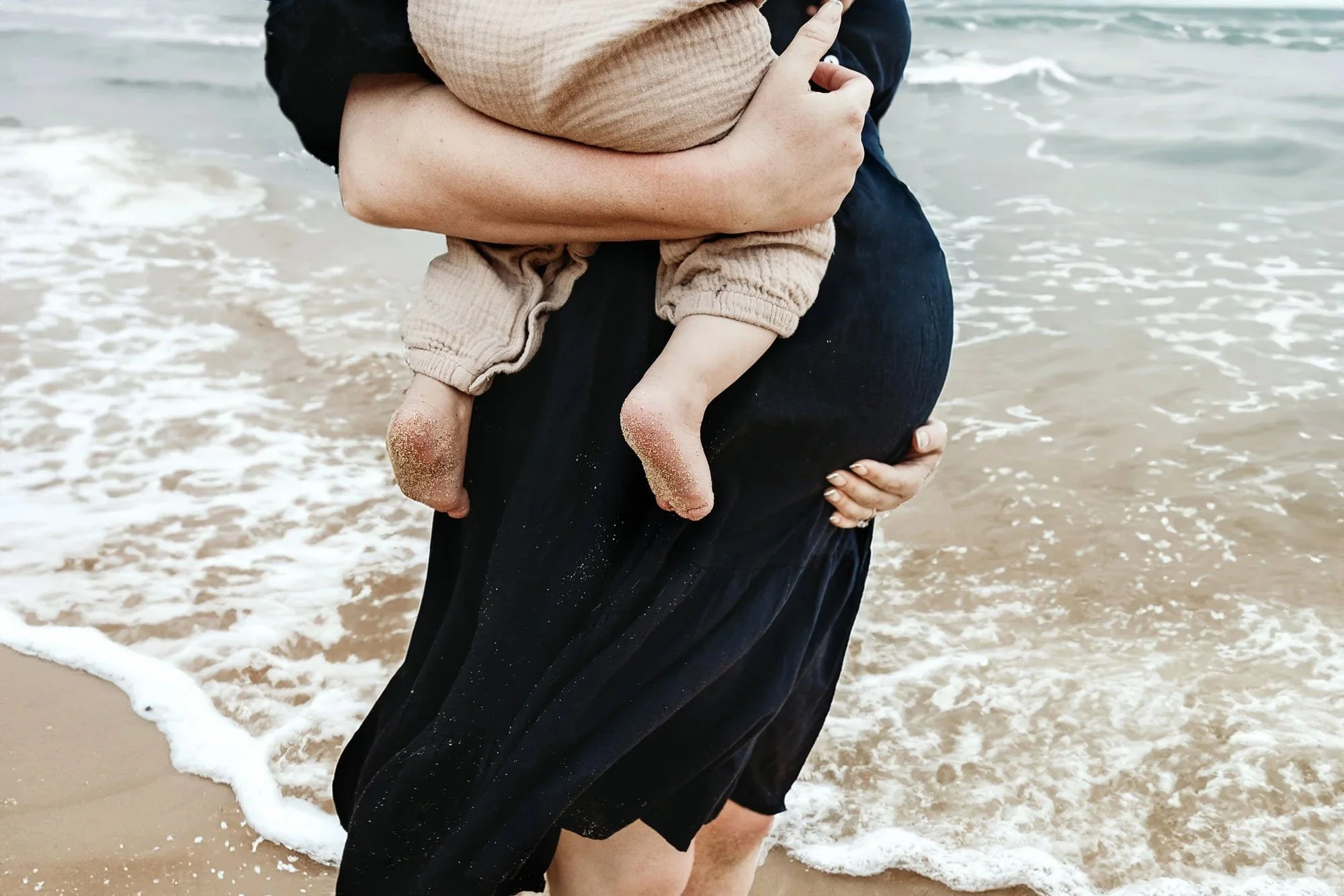 Pregnant mama holding baby bump on beach with daddy standing behind her kissing her