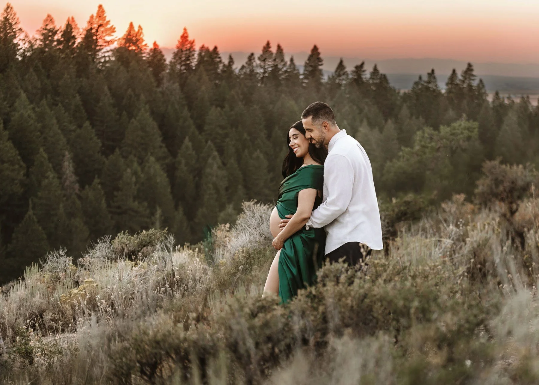 Couple in mountains with pine trees and sunset behind, baby bump visible