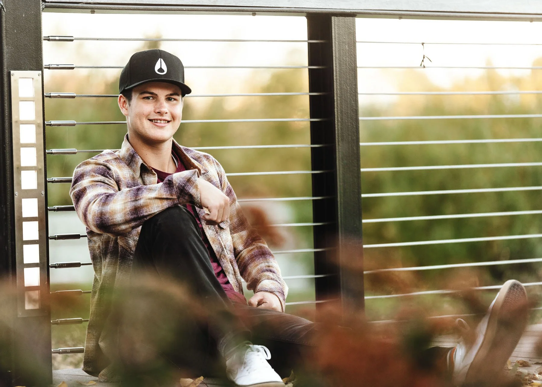 Teen boy in plaid shirt sitting in front of fence for senior session