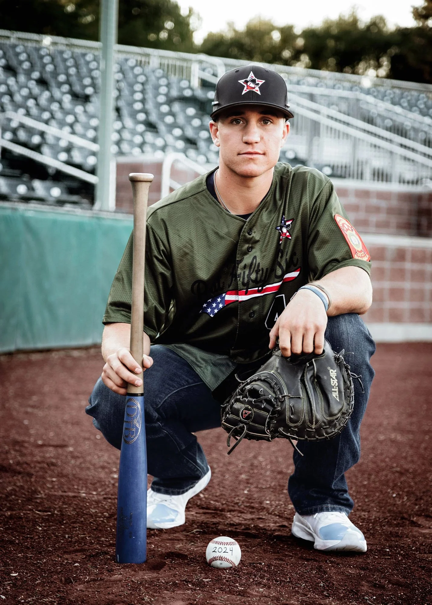 Senior boy squatting with mitt, baseball, and bat in front of baseball bleachers