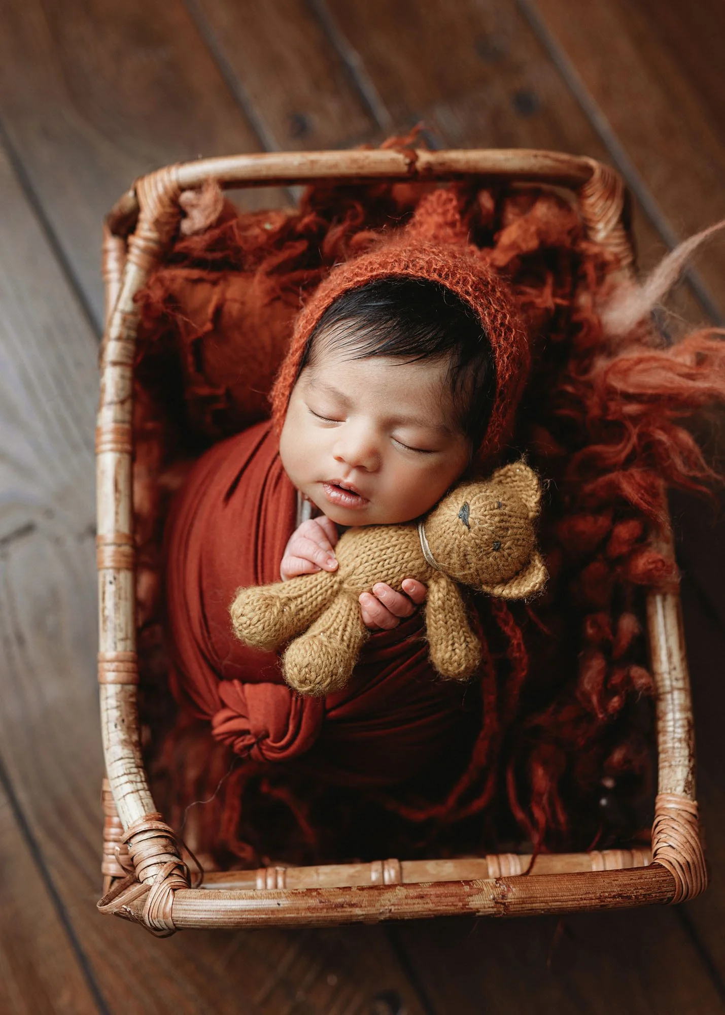 Newborn wrapped in rust orange swaddle holding miniature teddy bear