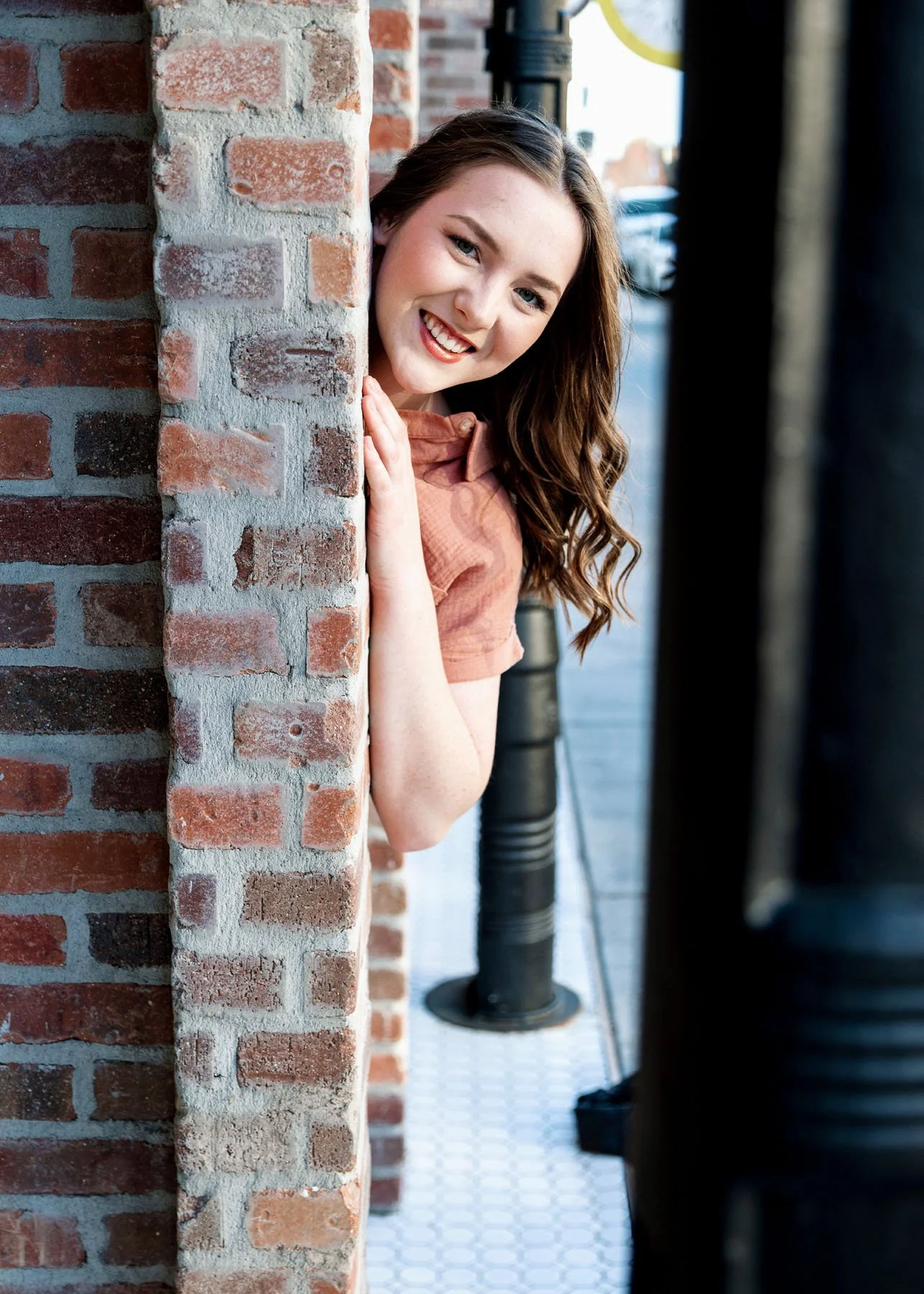 Senior girl peeking from behind brick wall in orange shirt