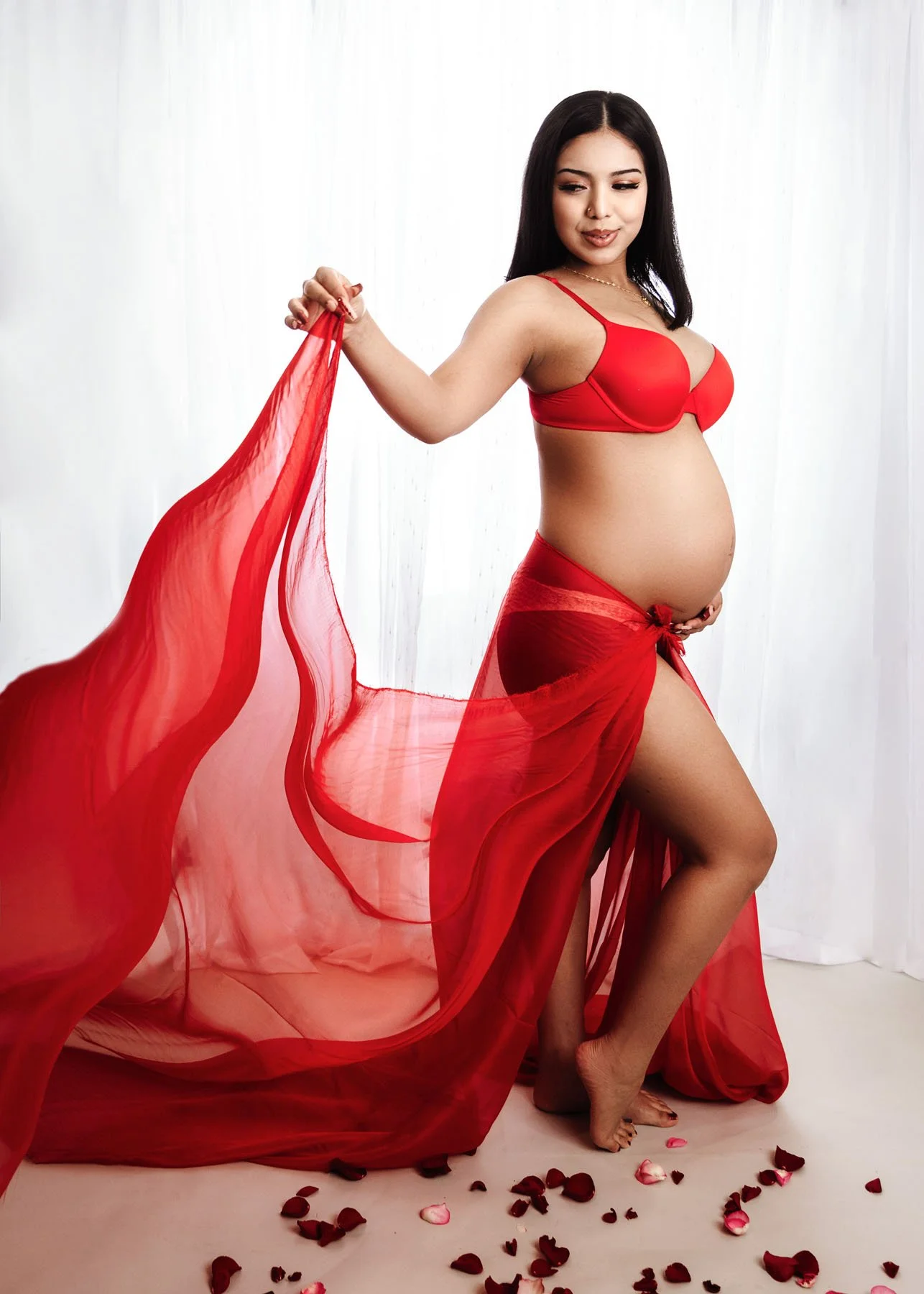 Third mama in red dress outdoors holding baby bump