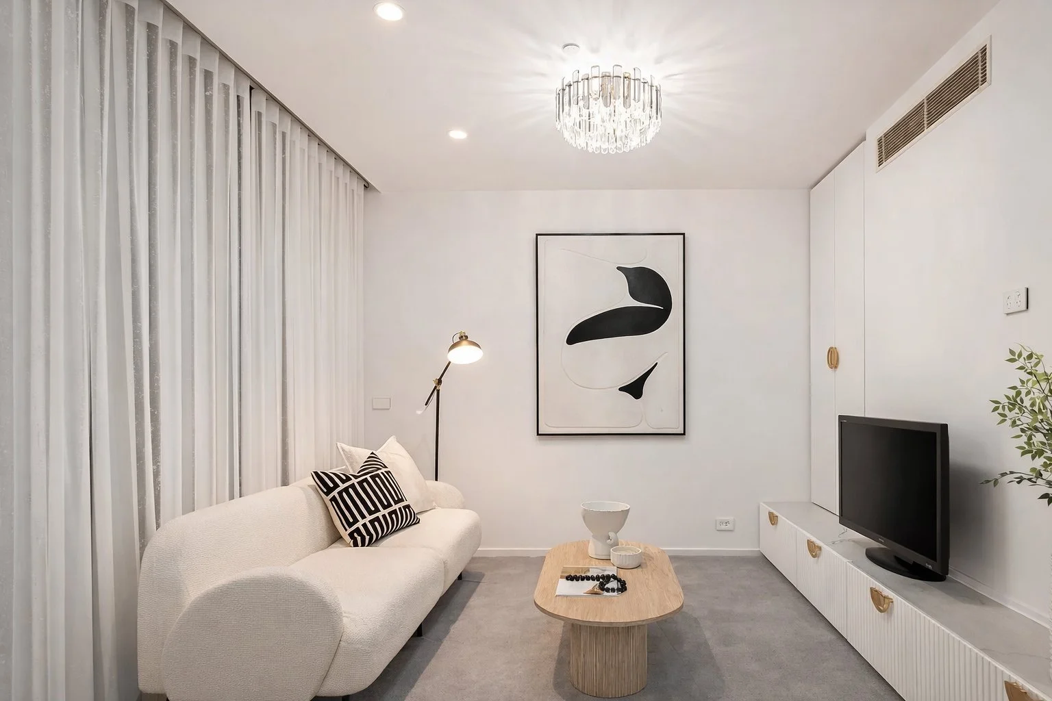 Modern living room with gray sofas, decorative vases, a round coffee table, and a television. Open kitchen and dining area with wooden furniture and sliding glass door leading outside. Bright with recessed ceiling lights.