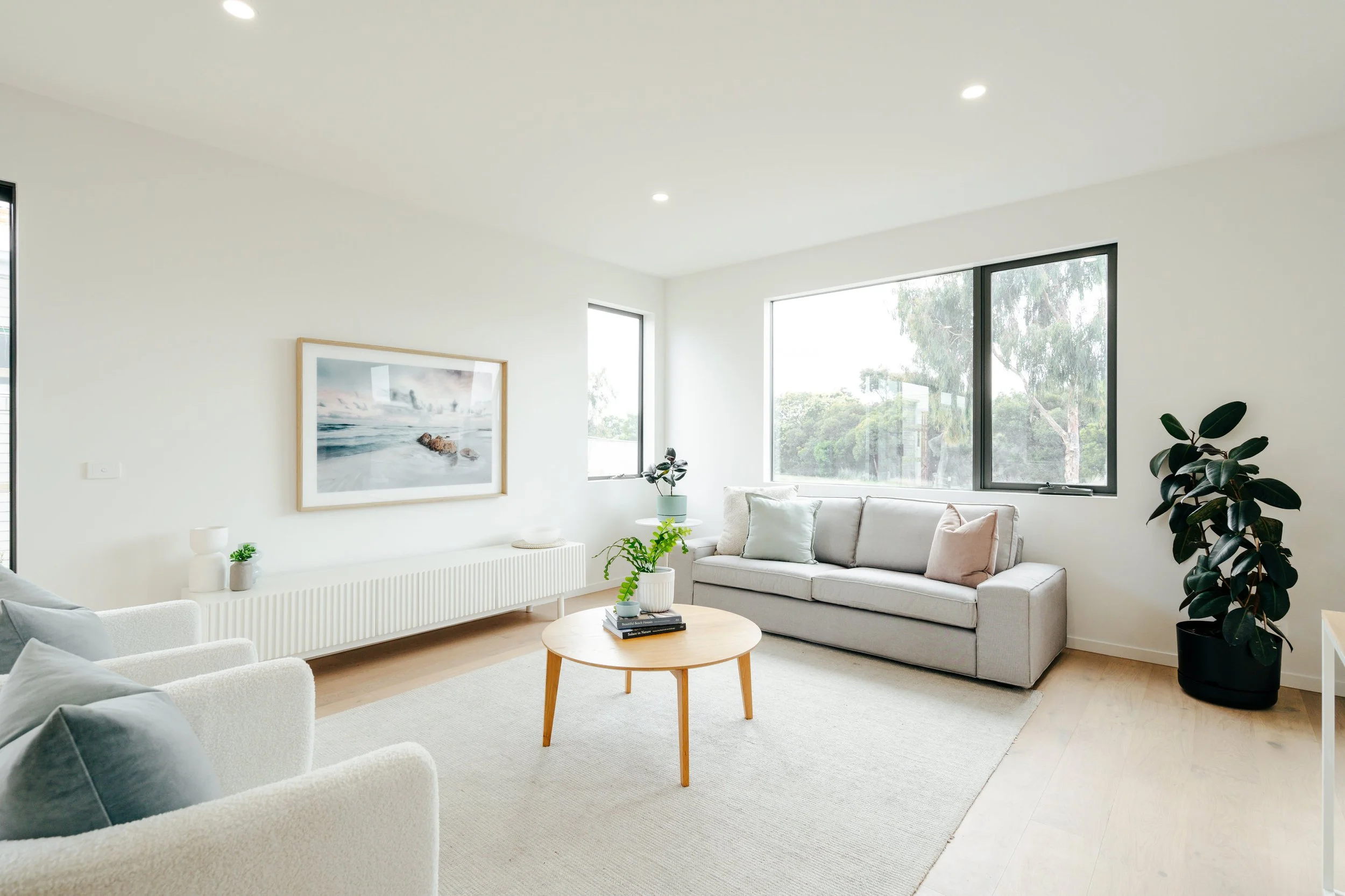 Bright living room with large windows, white walls, white sofa, armchair, light wooden table with plants, minimal decor, and a large painting of a seascape.