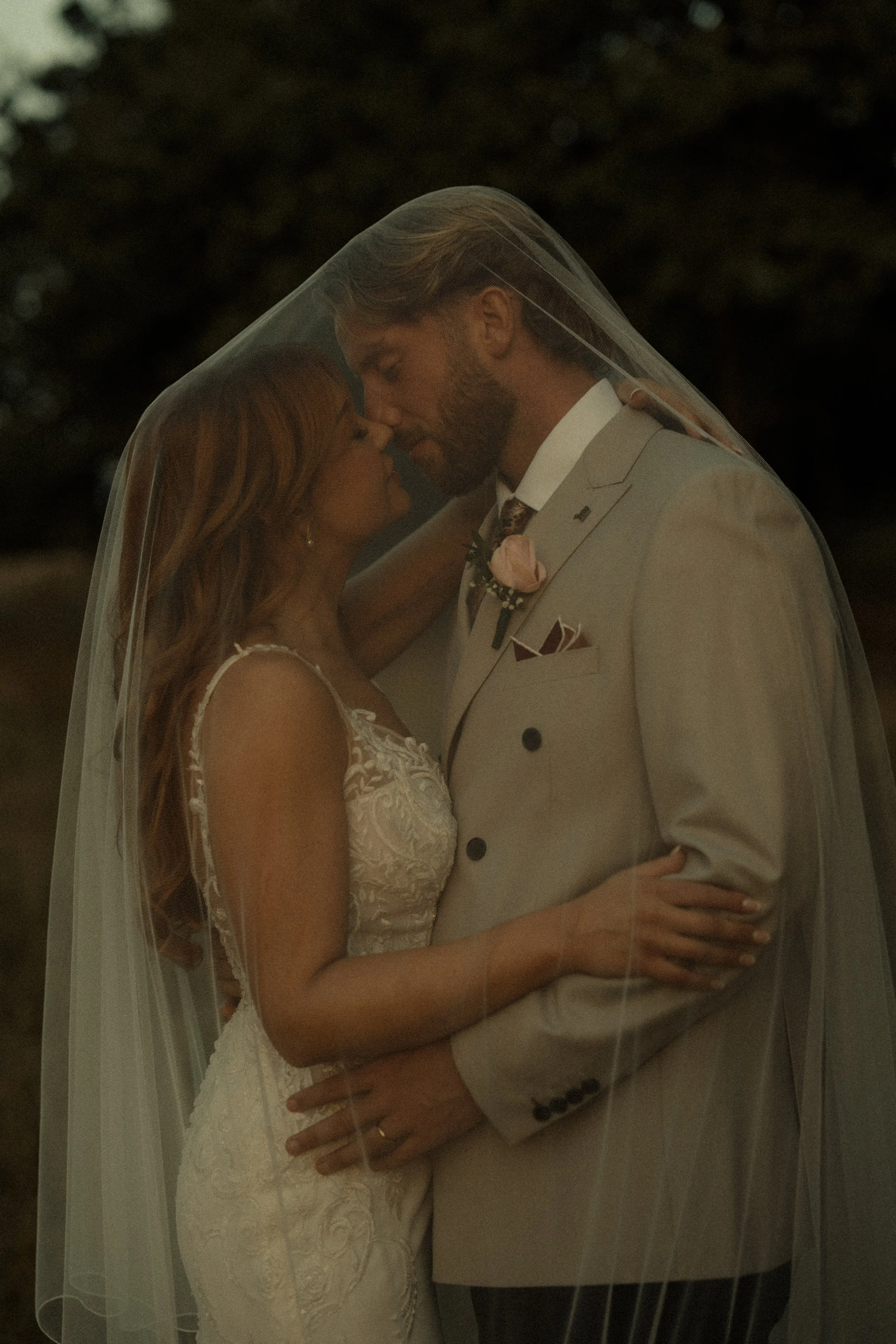 A bride and groom are close together, touching foreheads beneath a veil during their wedding, with dark background.