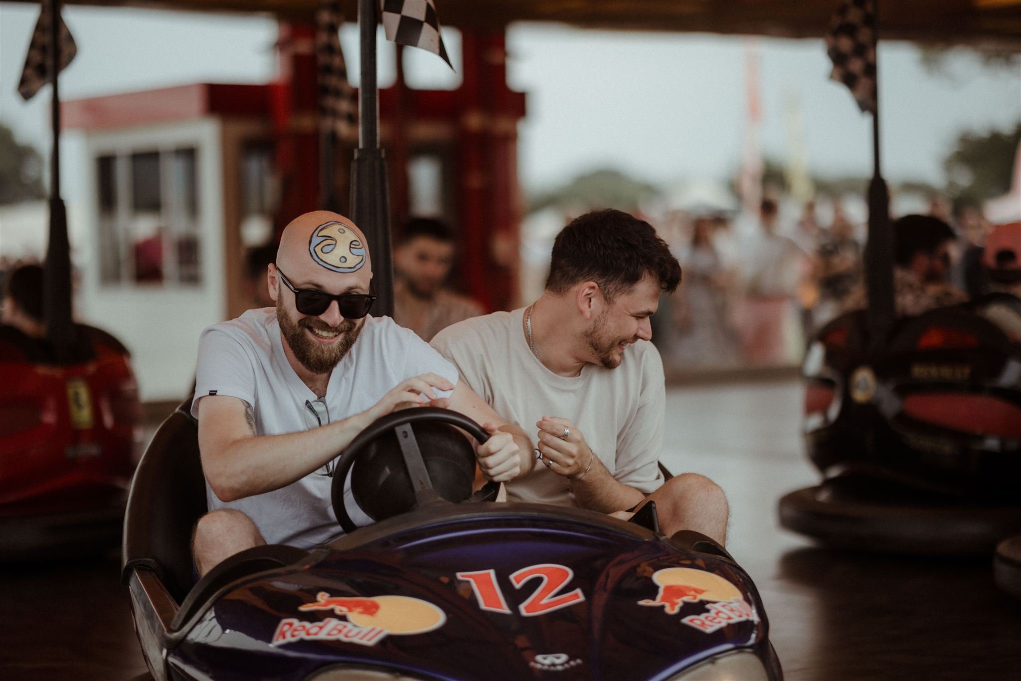 Two men laughing while riding a bumper car at an amusement park. One wears glasses and has a design painted on his head. The car is labeled with a Red Bull logo.