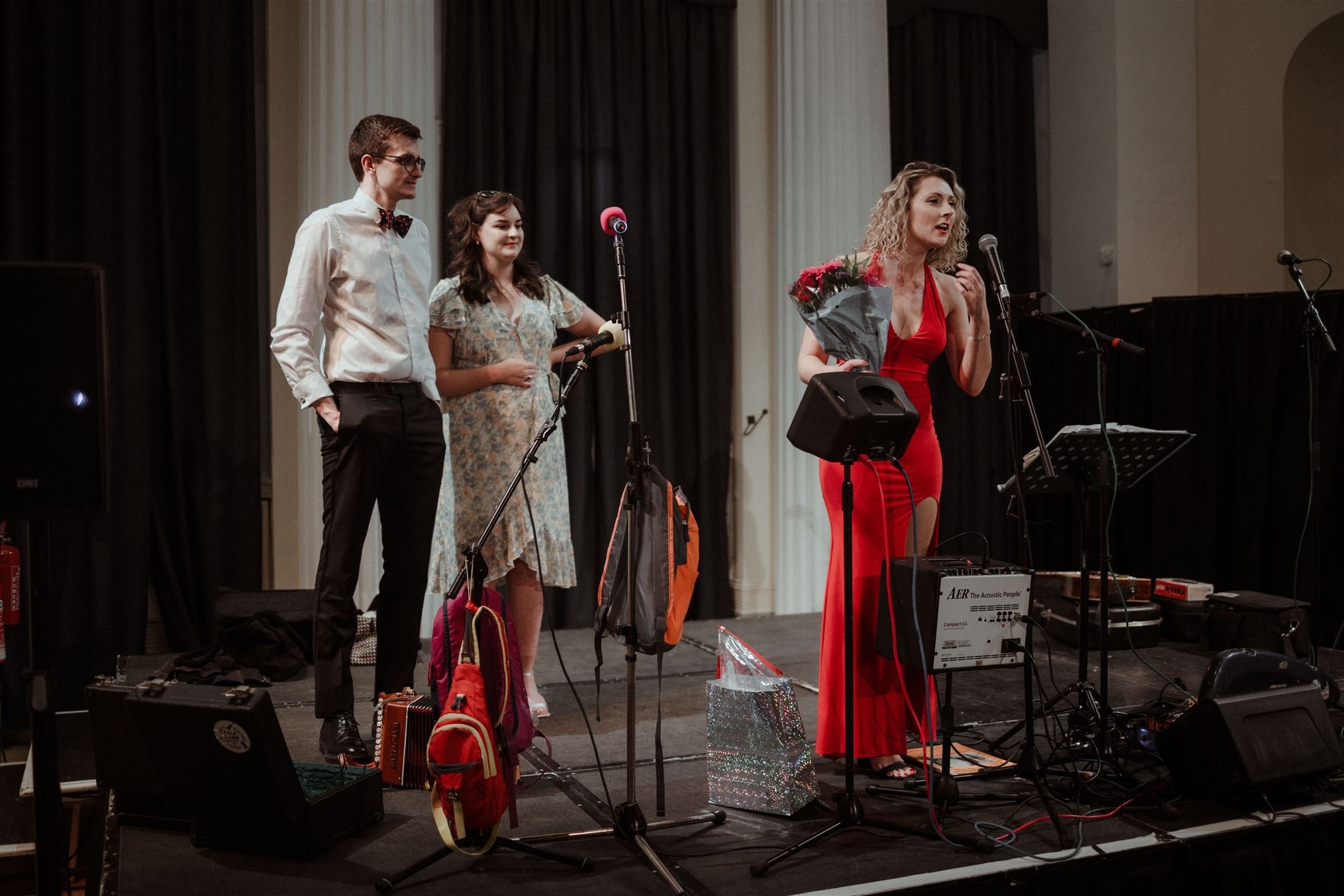 Three people on stage; one woman in a red dress holding flowers and speaking into a microphone, another woman in a floral dress, and a man in a white shirt and bow tie. Microphones and musical equipment are on stage.