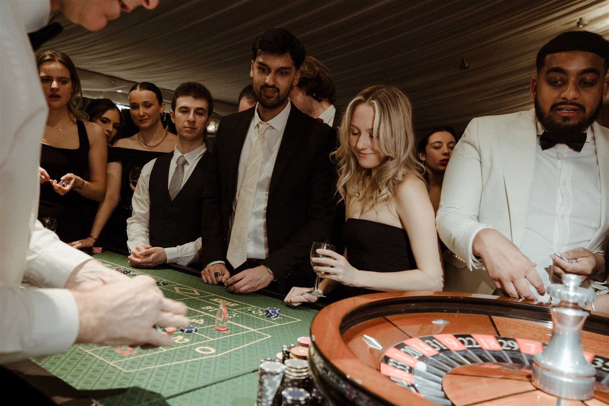 People gathered around a roulette table at a casino event, dressed in formal attire, engaging in gameplay and watching the dealer.