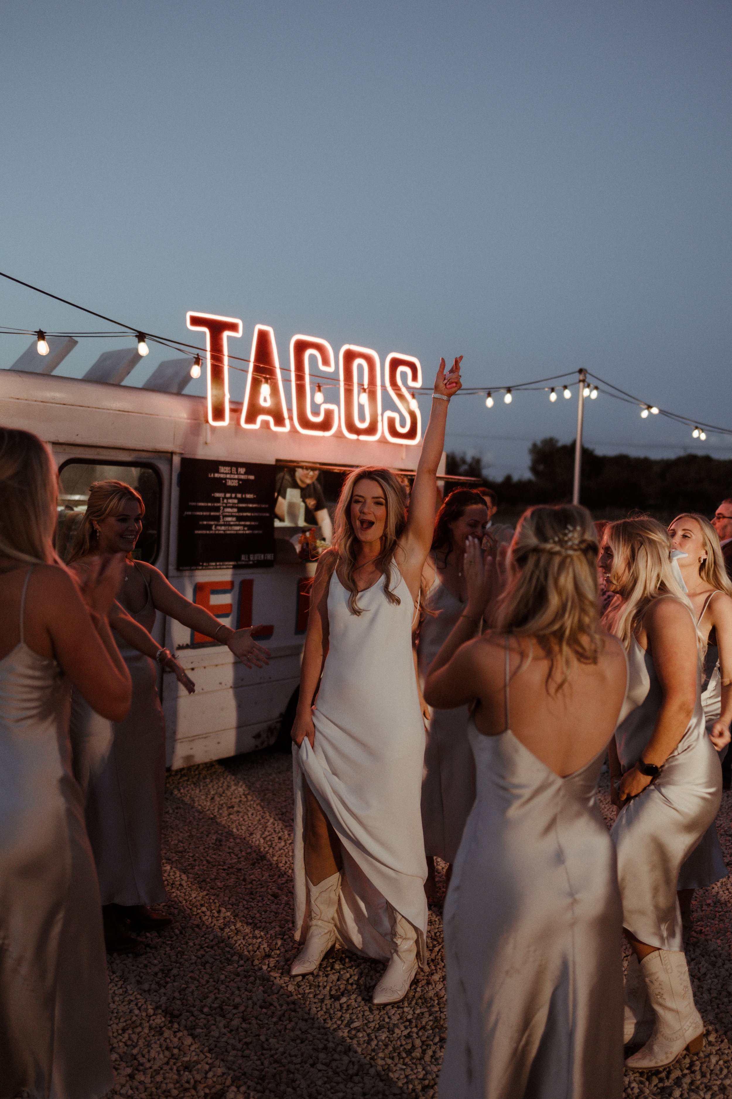 A group of women in white dresses celebrating near a taco truck with a neon "Tacos" sign at dusk.