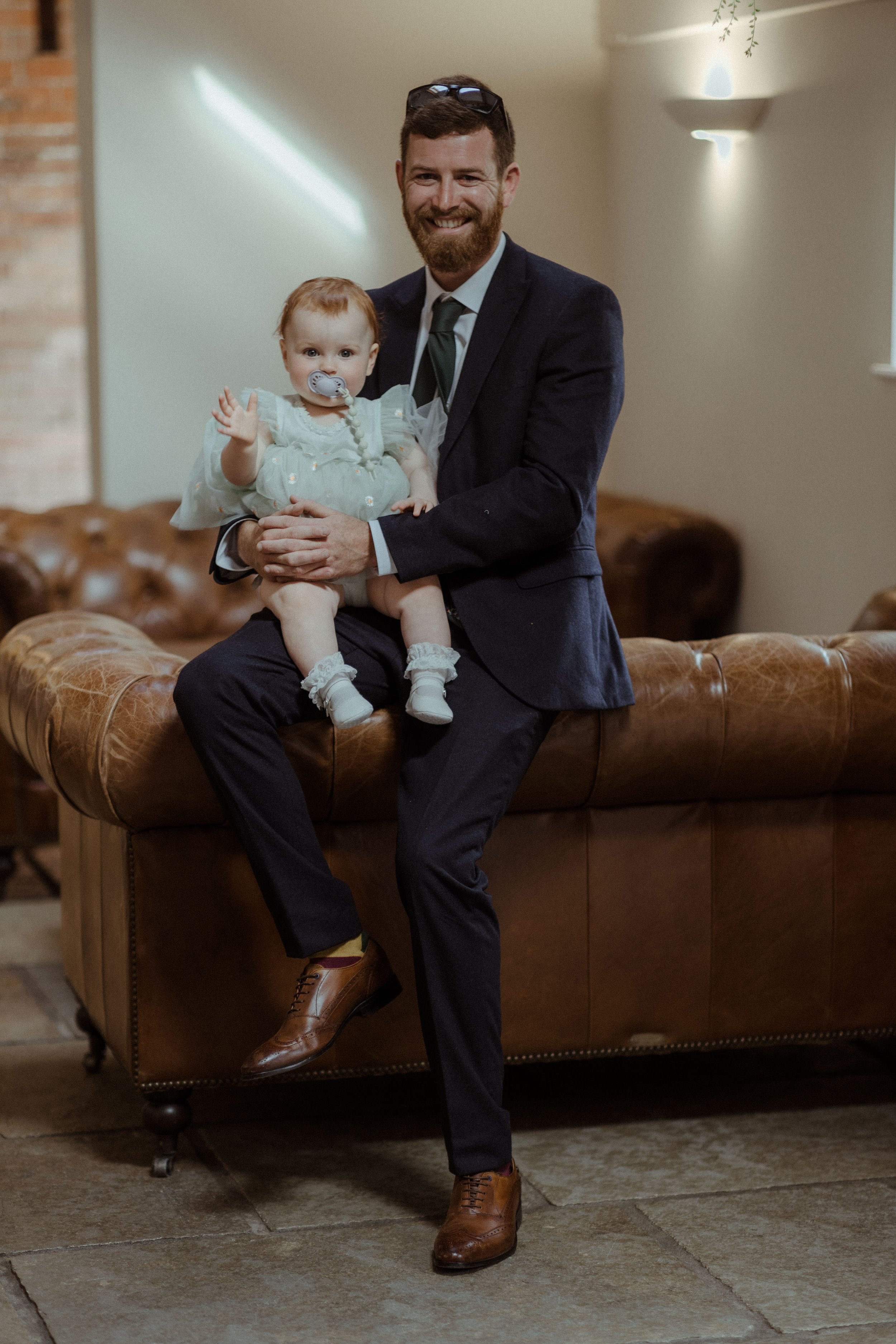 Man in a suit sitting on a brown leather couch holding a baby wearing a light blue dress and pacifier.
