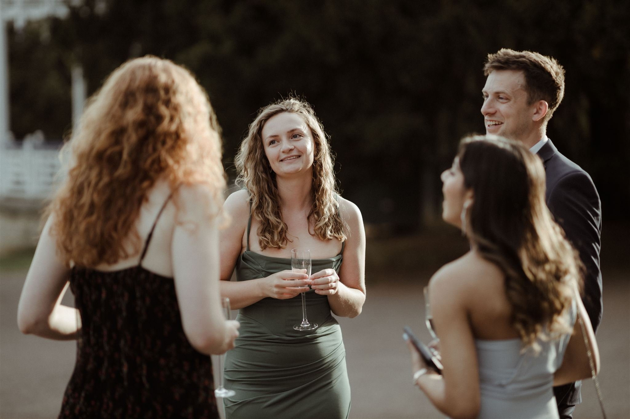 A group of four people socializing outdoors, dressed in formal attire and holding drinks.
