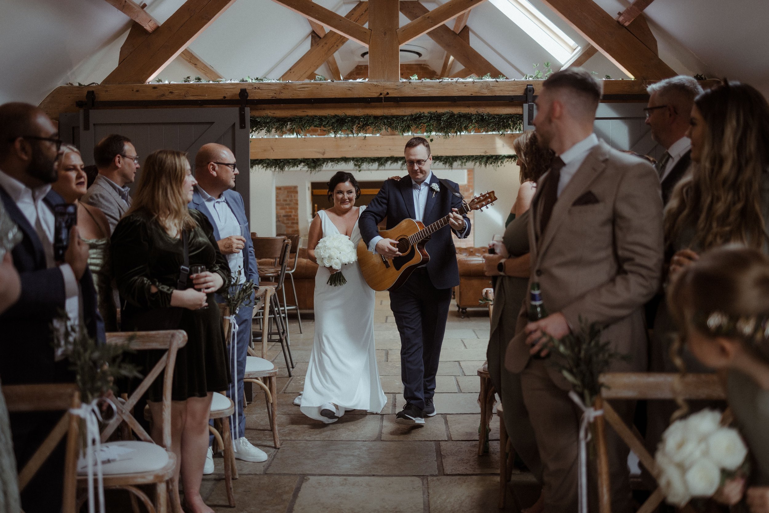 Bride and groom walking down an aisle while groom plays guitar at a wedding ceremony, surrounded by guests in a rustic venue.