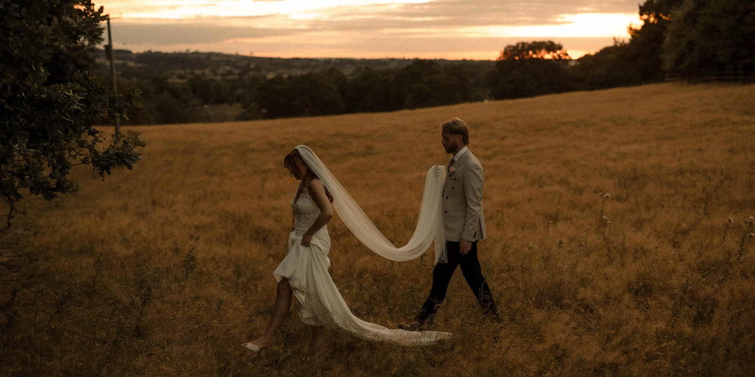 A bride and groom walking through a field during sunset, with the bride wearing a wedding dress with a long train and veil, and the groom in a light suit.