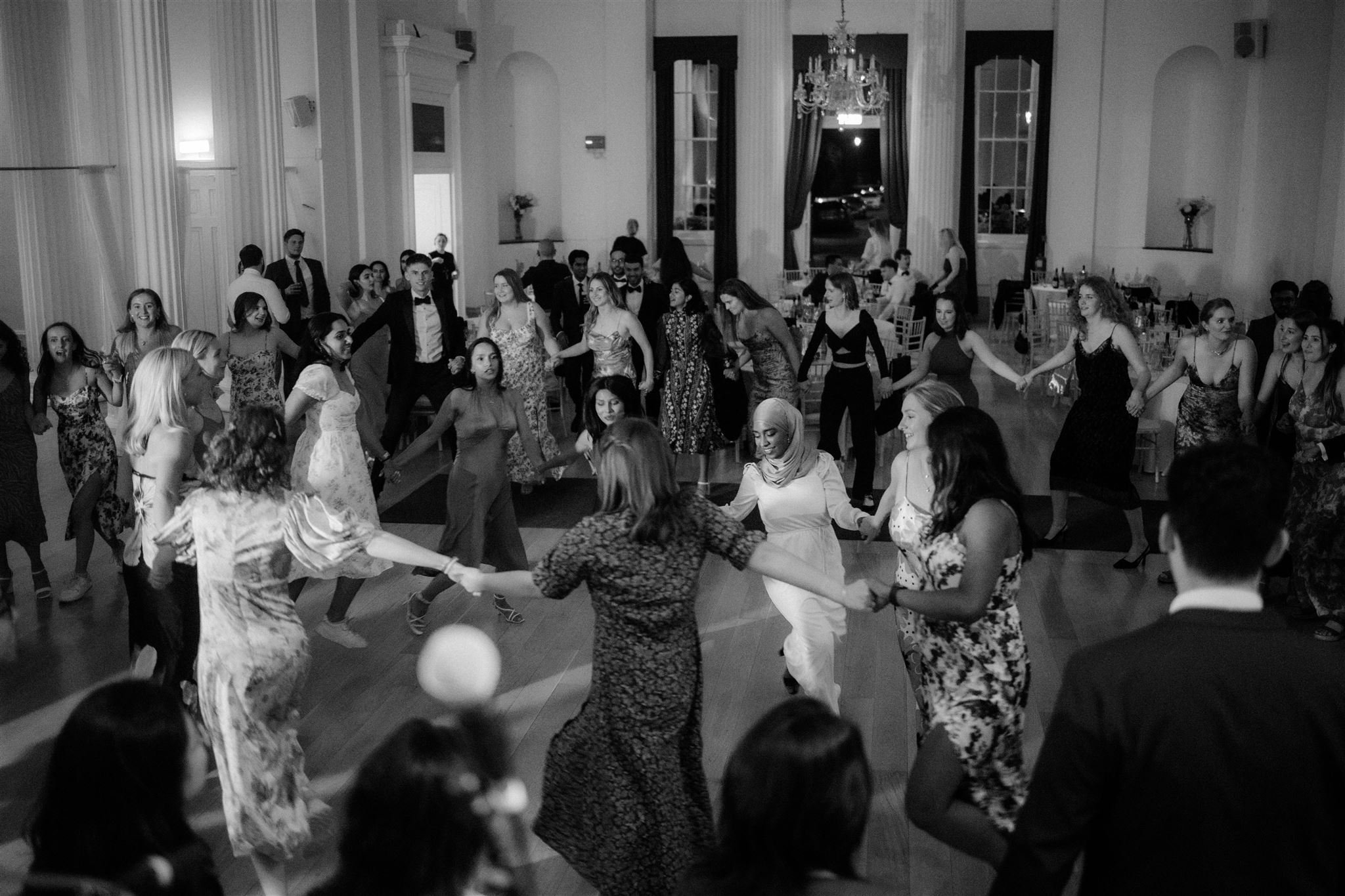 Group of people holding hands and dancing in a circle at a formal event in an elegant room with large windows and a chandelier.