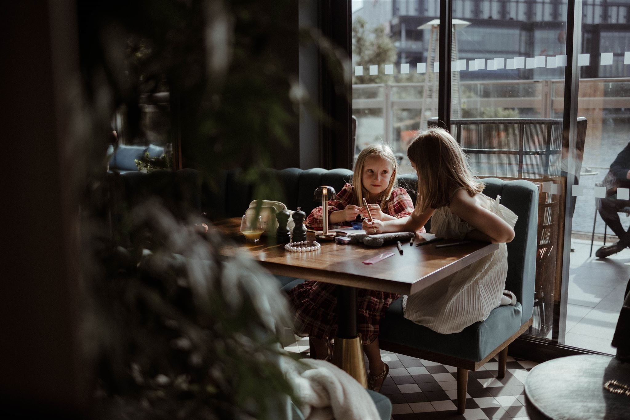 Two young girls sitting at a table in a cafe, drawing with colored pencils on paper, with large windows in the background letting in natural light.