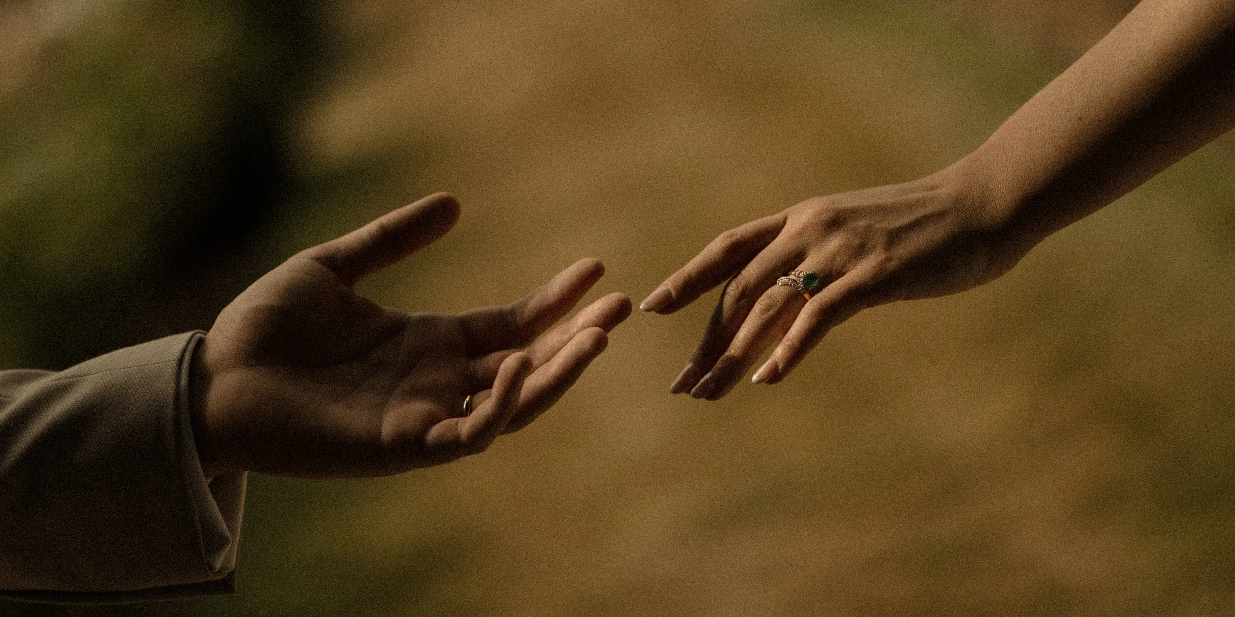 Two hands reaching towards each other, one with a ring, set against a blurred background.