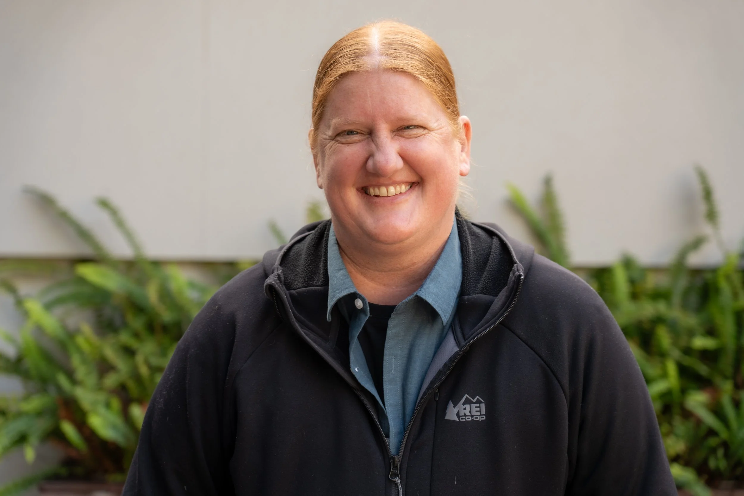 Headshot of Jen Walsh, smiling and standing in front of green shrubs