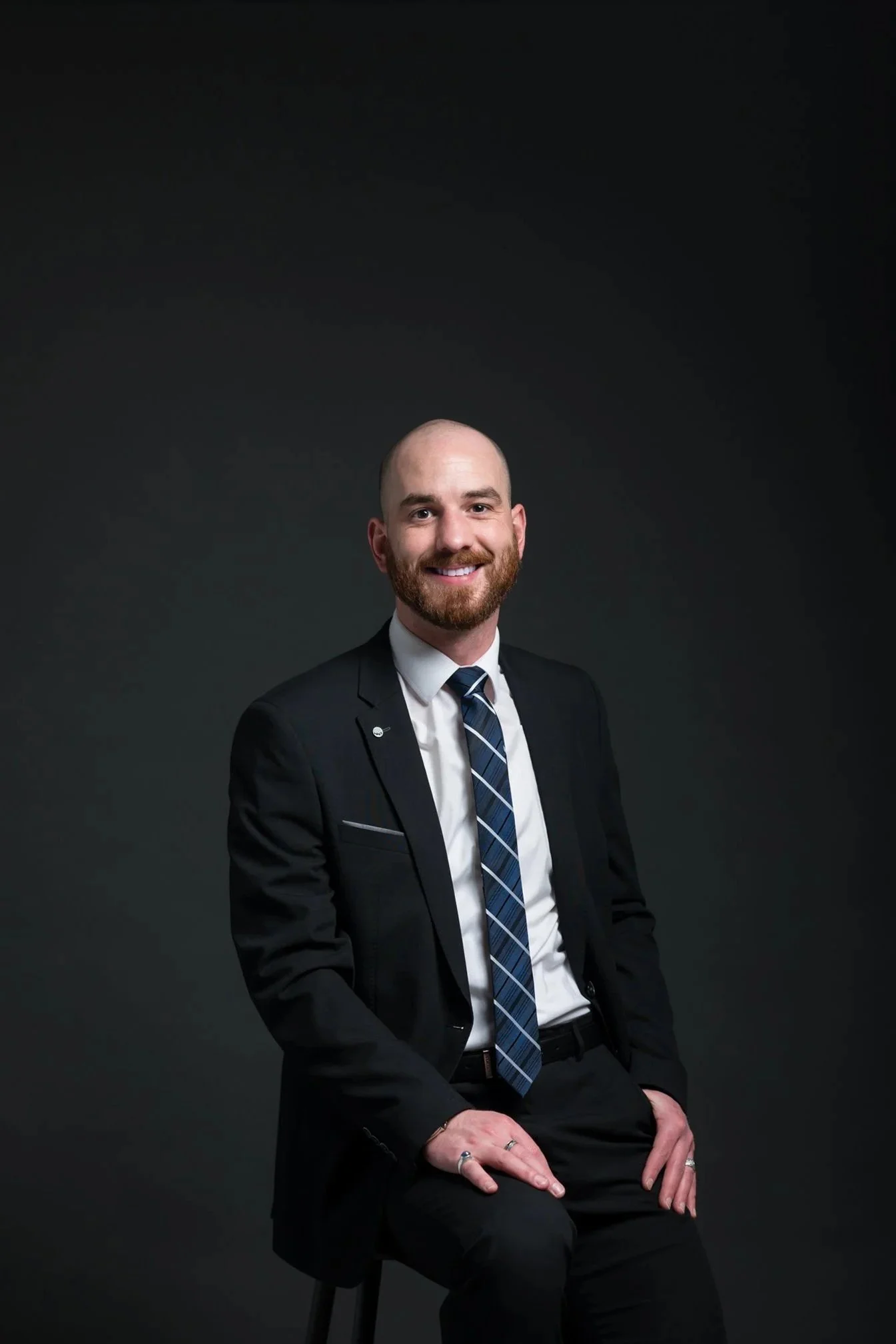 A smiling man in a black suit, white shirt, and striped tie, sitting against a dark background.